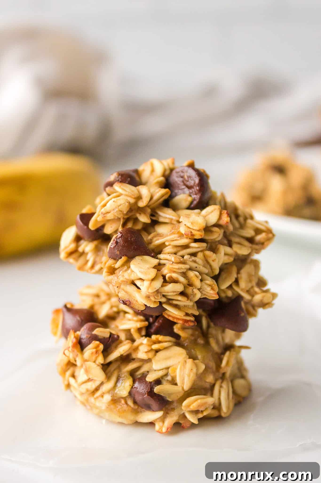 Two golden-brown banana oatmeal cookies, subtly studded with chocolate chips, are artfully stacked on a clean white surface, showcasing their delicious texture.