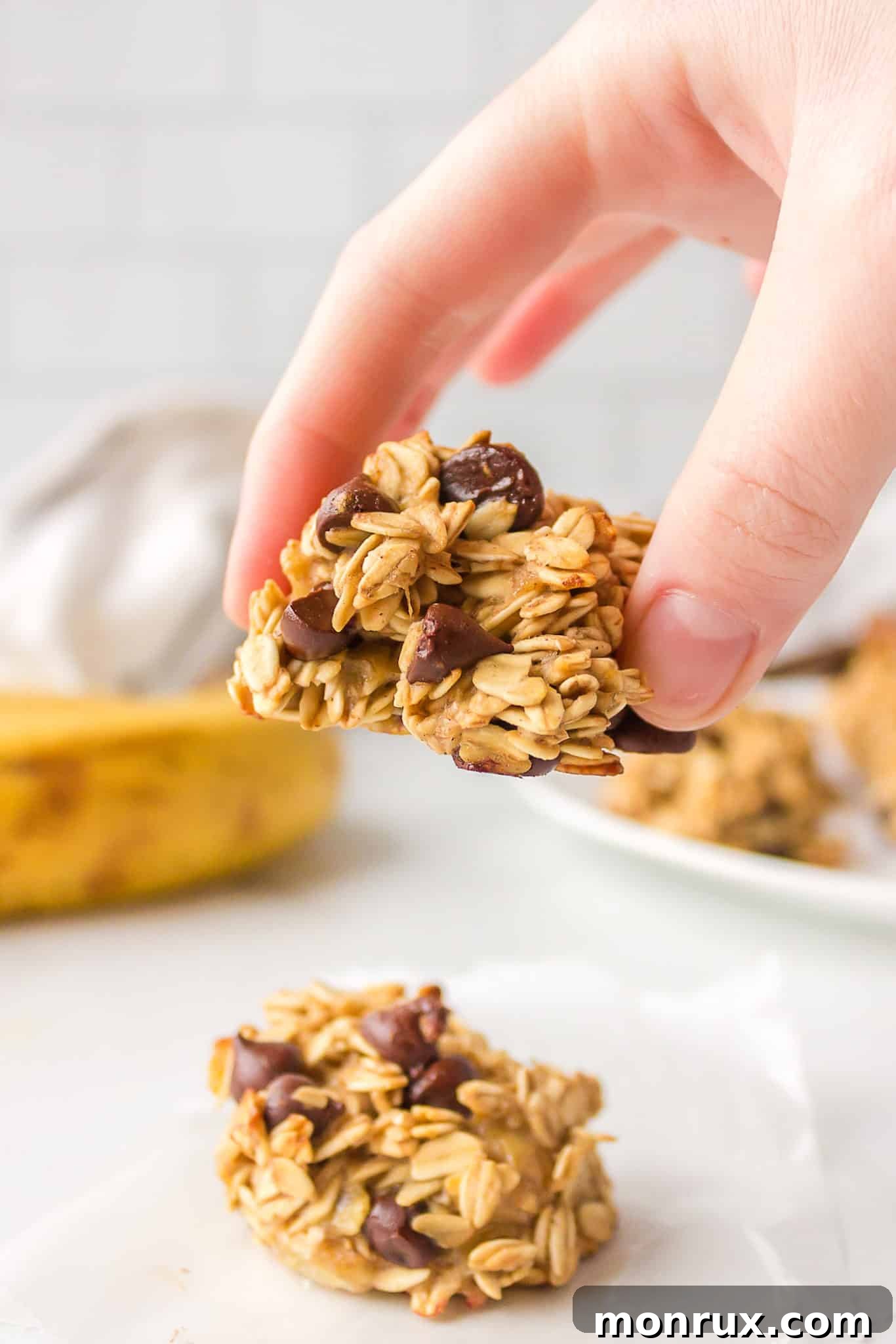 A hand gently holds a freshly baked, vegan banana oatmeal cookie with visible chocolate chips, highlighting its soft texture and delicious appeal.