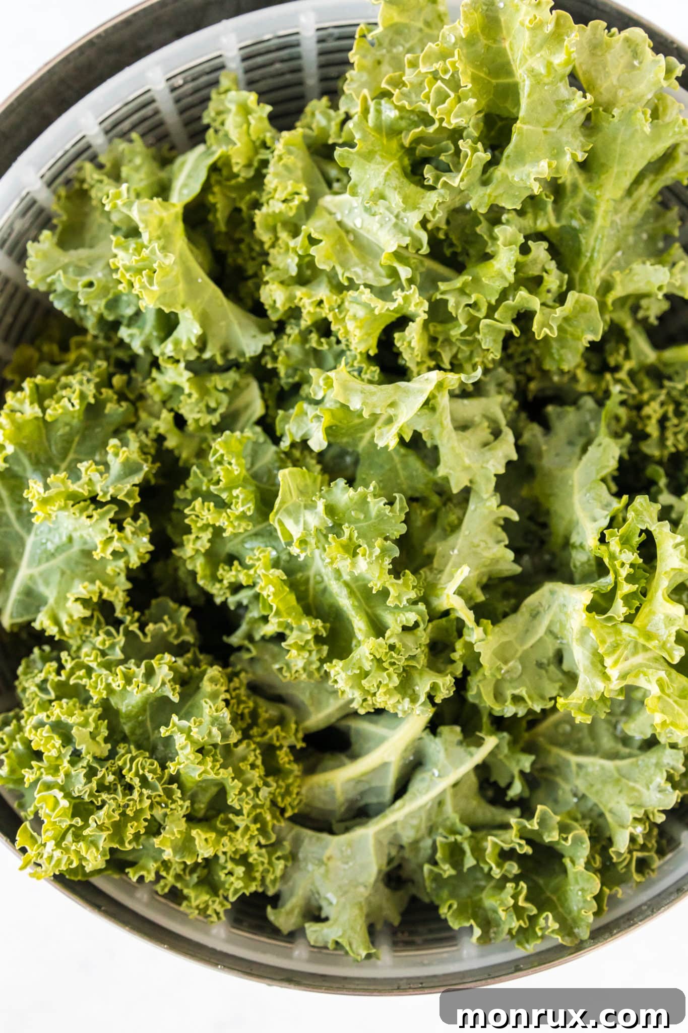 Fresh curly kale leaves are shown rinsing under water in a metal colander, preparing them for baking.