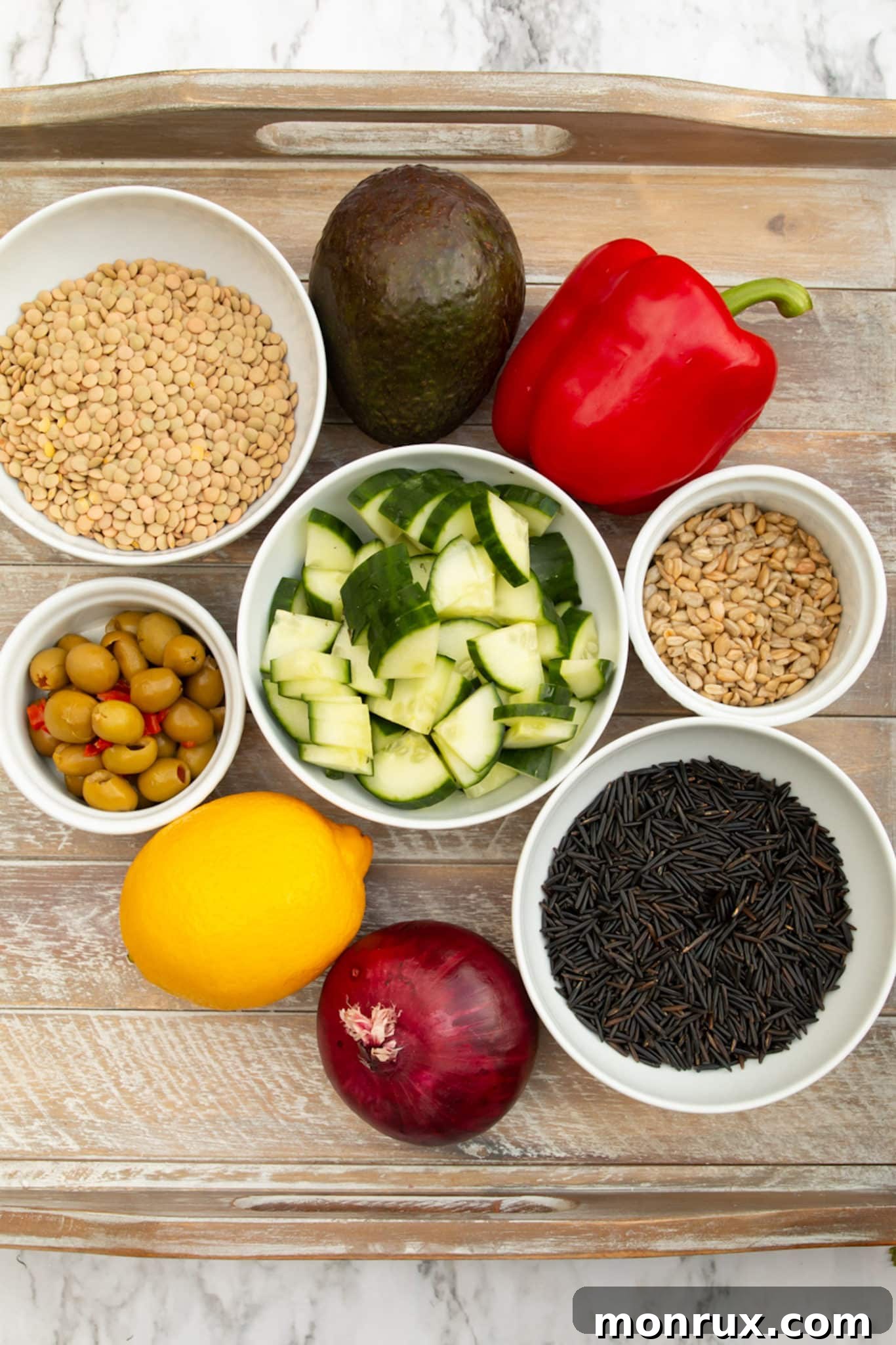 A flat lay photograph of various ingredients for wild rice and lentil salad including cooked grains, fresh vegetables, olives, lemon, and sunflower seeds.