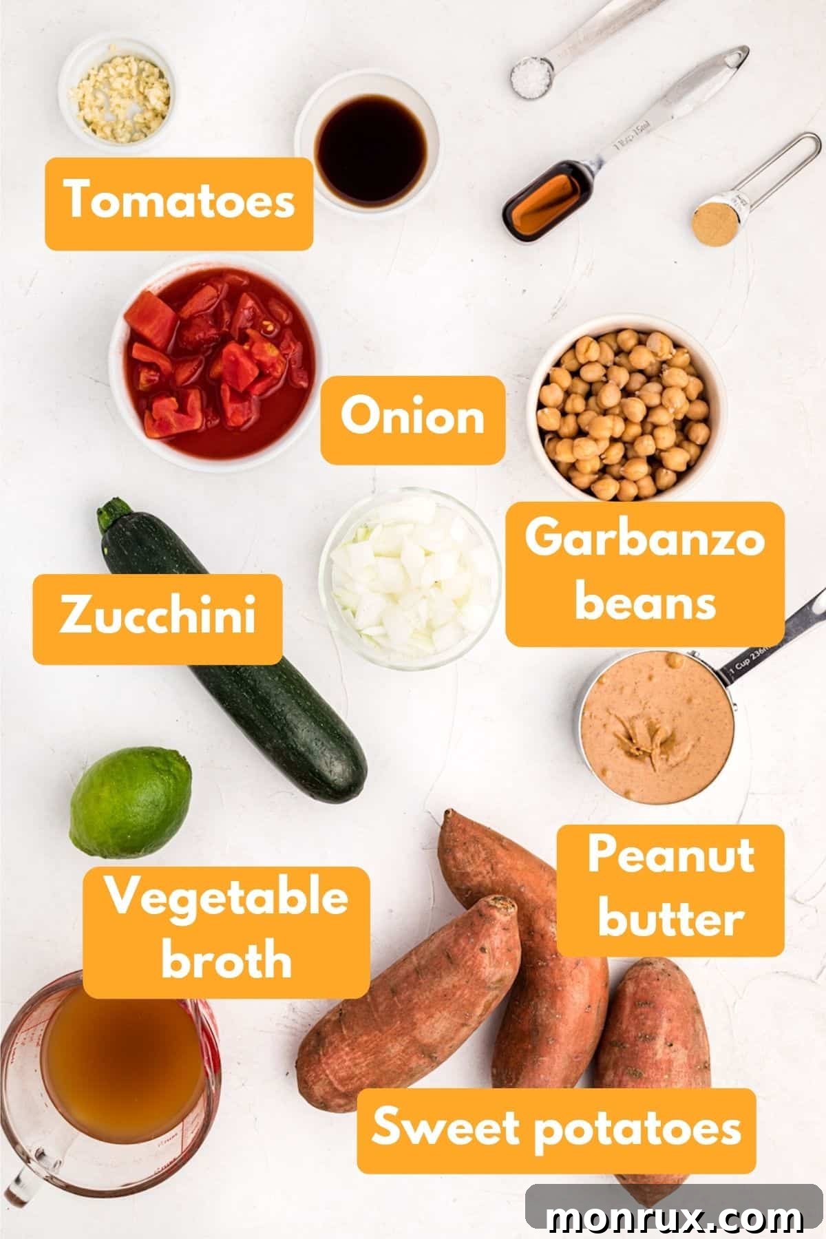A vibrant collection of fresh ingredients for peanut butter soup, including sweet potatoes, zucchini, onions, tomatoes, garbanzo beans, and a jar of natural peanut butter, neatly arranged on a rustic wooden surface.
