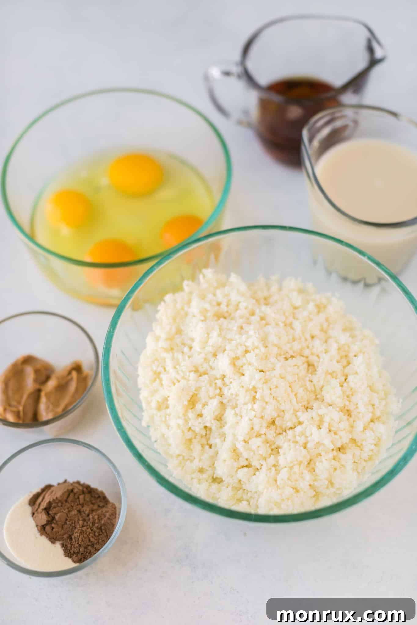 Assortment of key ingredients for cauliflower oatmeal laid out on a kitchen countertop, including cauliflower, eggs, almond milk, cocoa powder, almond butter, and collagen protein.