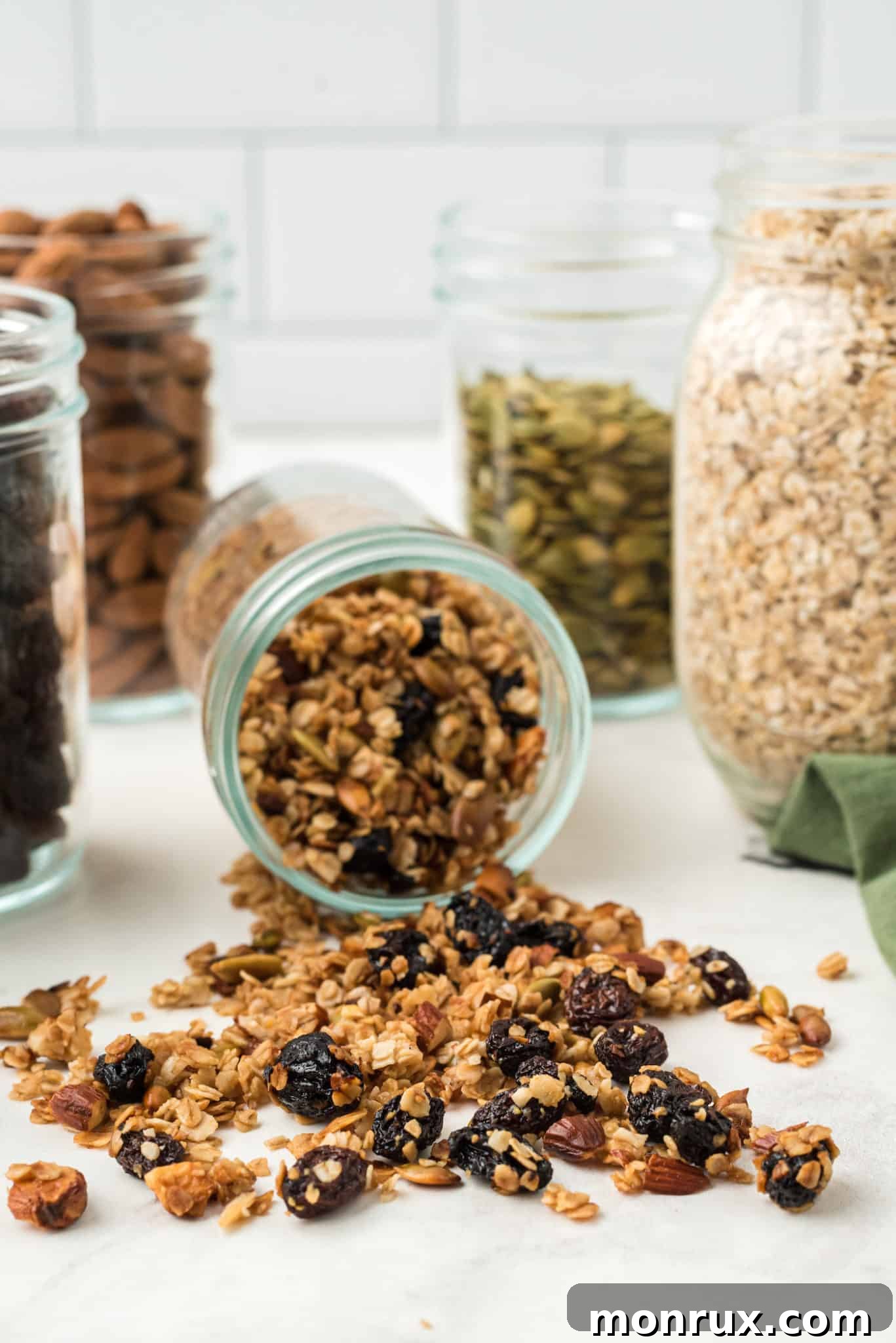 A glass jar of air fryer granola lying on its side, surrounded by scattered oats and dried fruit, highlighting its texture and homemade appeal.