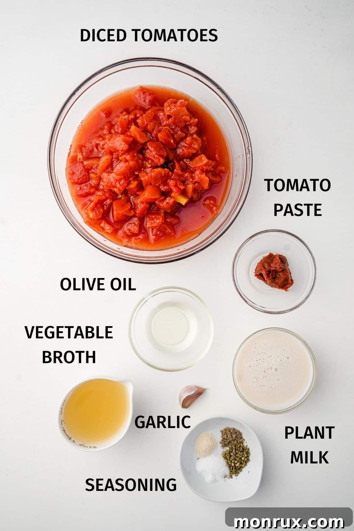 A flat lay image displaying the key ingredients for tomato soup, including canned diced tomatoes, vegetable broth, plant milk, tomato paste, garlic, and herbs.