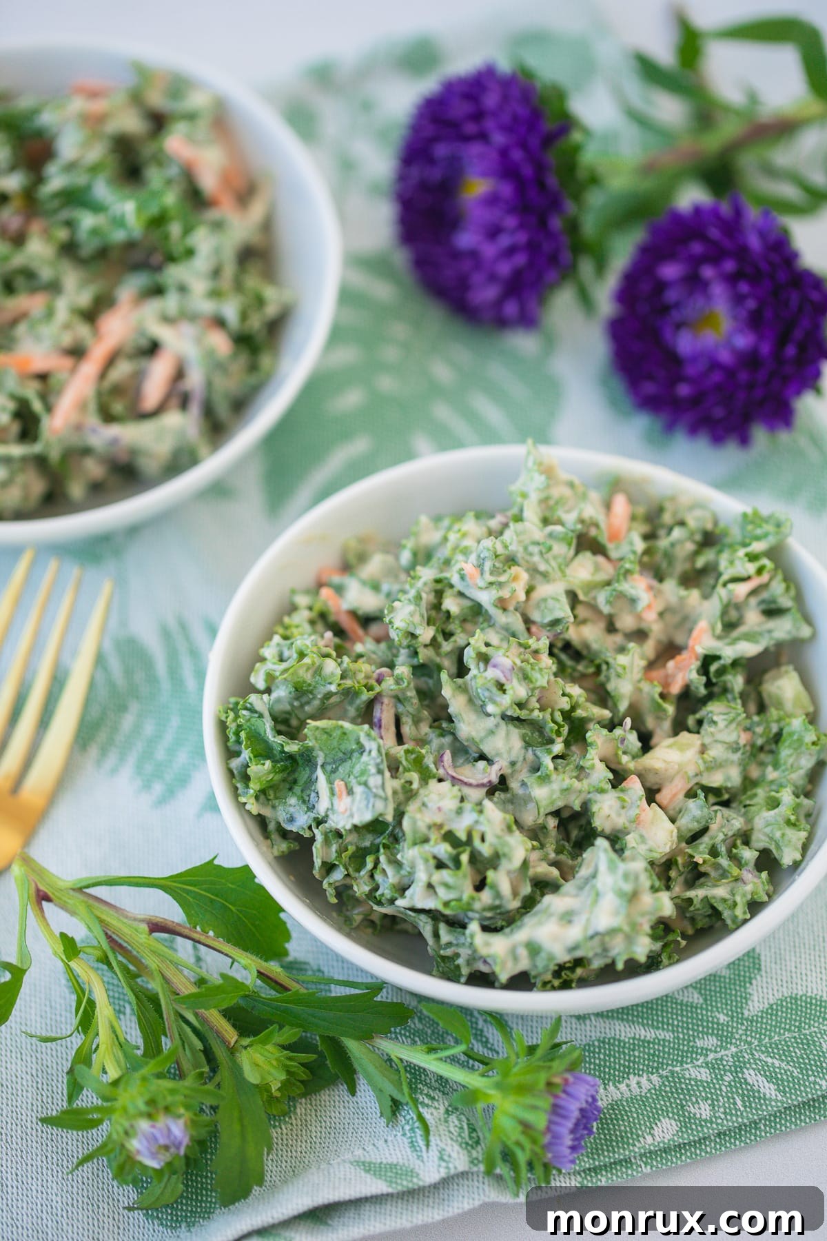 A beautifully arranged bowl of creamy kale salad resting on a green floral napkin, adorned with fresh purple flowers, highlighting its freshness and appeal.