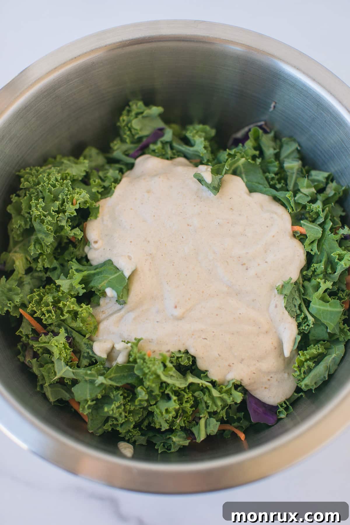 Shredded kale in a bowl with the creamy dressing poured generously on top, awaiting mixing.
