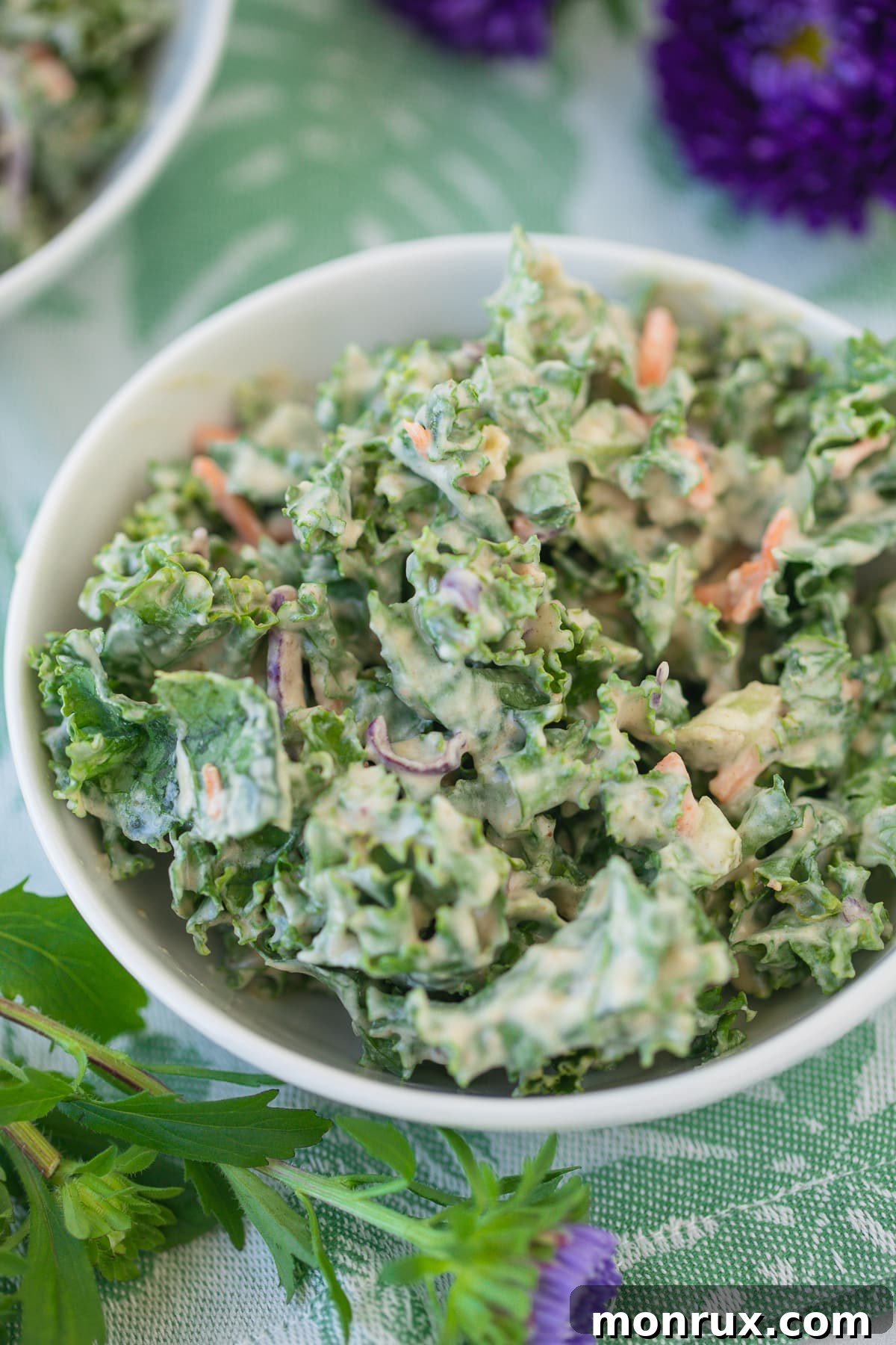 A beautifully presented bowl of creamy kale salad, adorned with purple flowers on a table, signifying a finished and ready-to-eat dish.