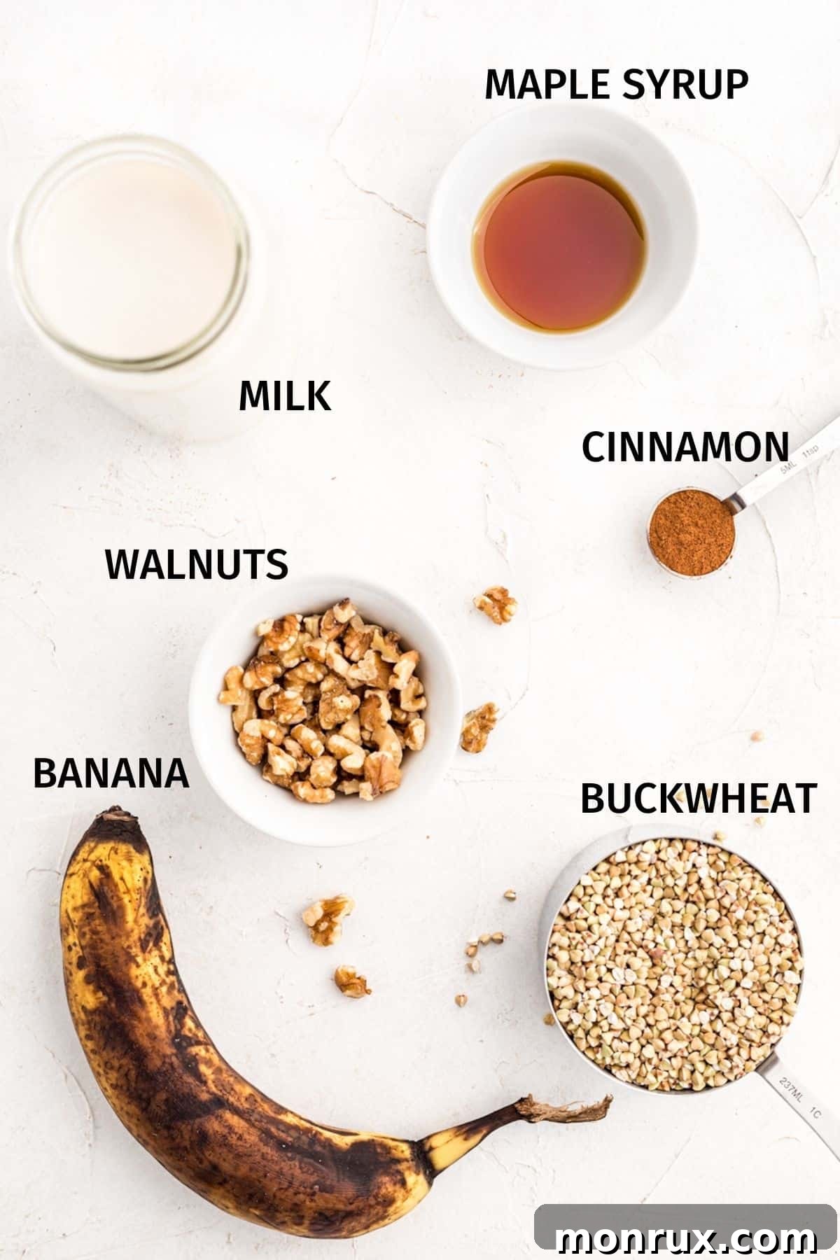 Ingredients for buckwheat porridge in small bowls on a white surface, including buckwheat groats, almond milk, maple syrup, banana, and walnuts.