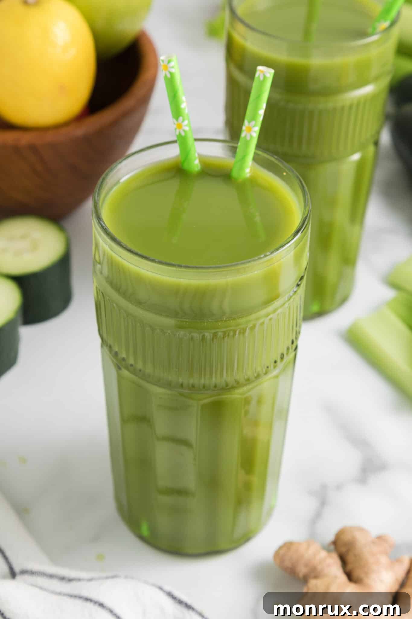 A tall glass of vibrant green juice, garnished with lemon slices and fresh mint, with two straws. The juice reflects light beautifully, emphasizing its refreshing nature.