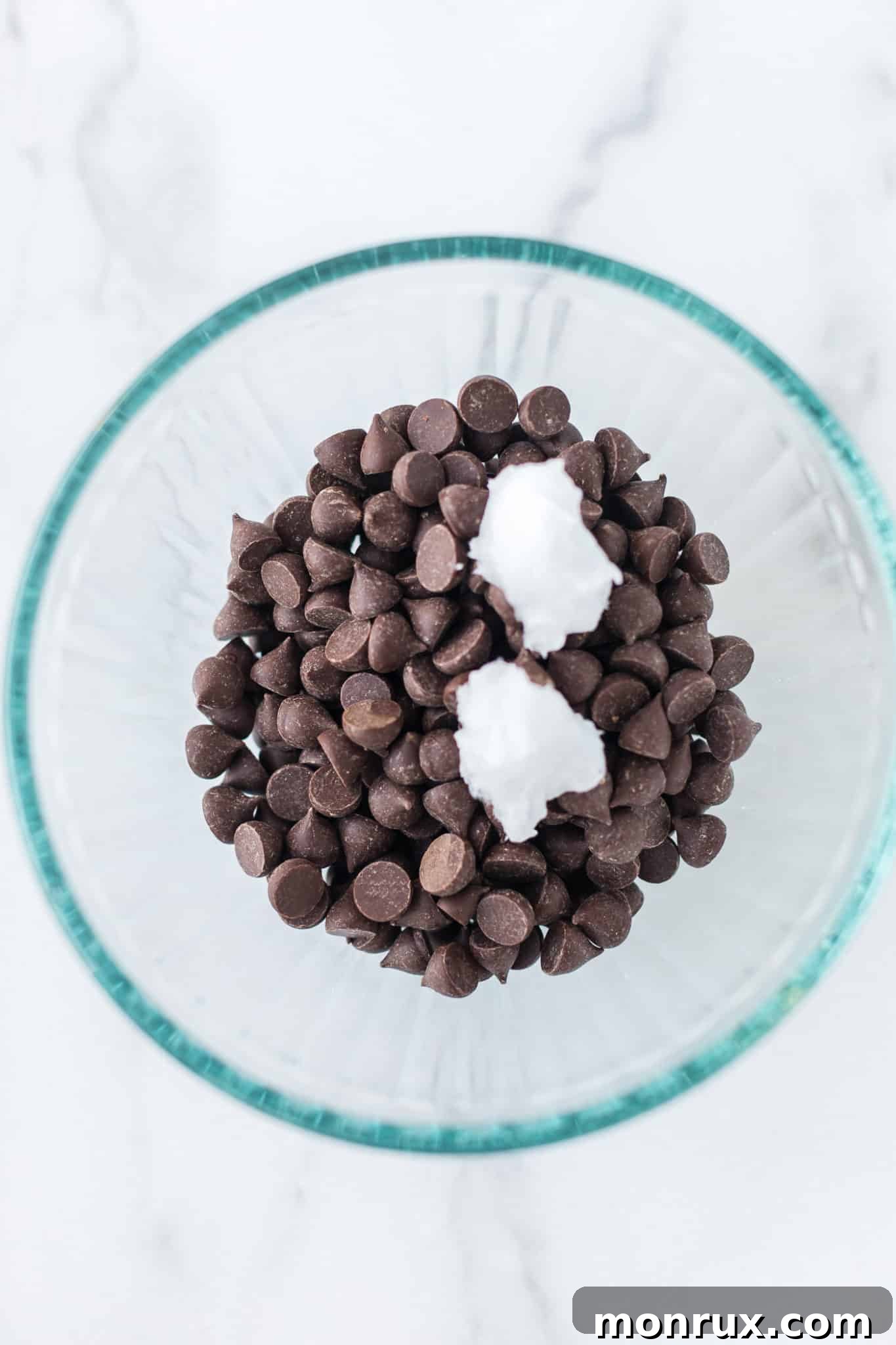 Unmelted dark chocolate chips and solid coconut oil placed together in a transparent glass bowl, perfectly arranged and ready for the melting process in the microwave.