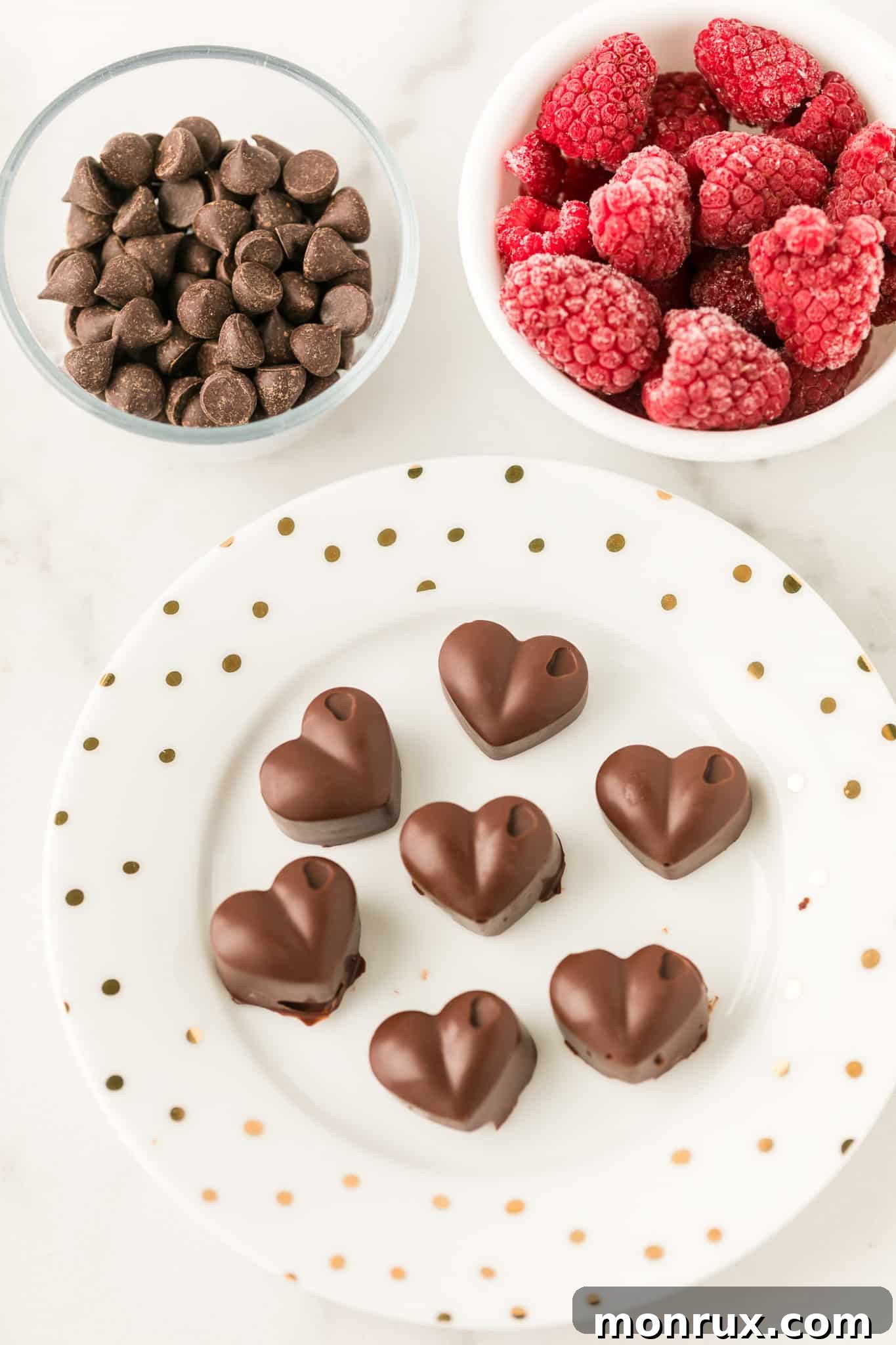 A beautiful arrangement of perfectly formed dark chocolate hearts presented on a white plate, positioned next to a bowl of chocolate chips and a separate bowl of vibrant frozen raspberries, indicating the ingredients and the delicious final product.