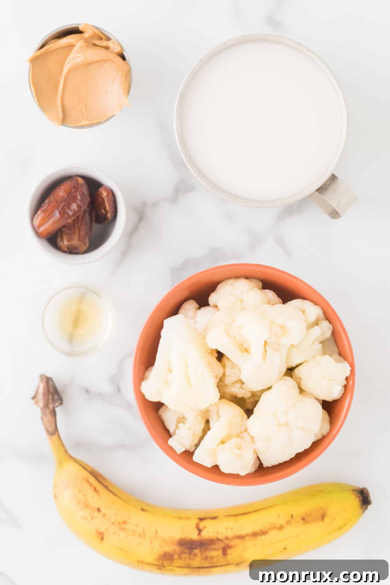 A selection of fresh and frozen ingredients including frozen cauliflower, Medjool dates, bananas, and a jar of natural peanut butter, neatly arranged on a counter.