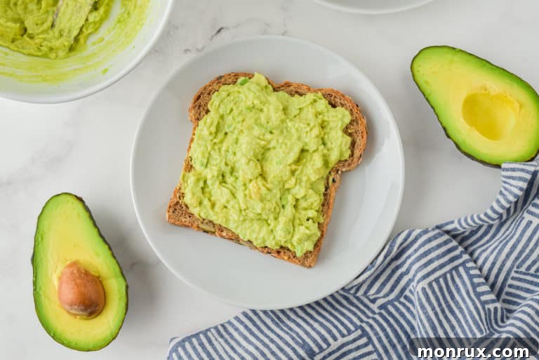 Top view of a white plate with a slice of avocado toast and a halved avocado, symbolizing a fresh and healthy meal.