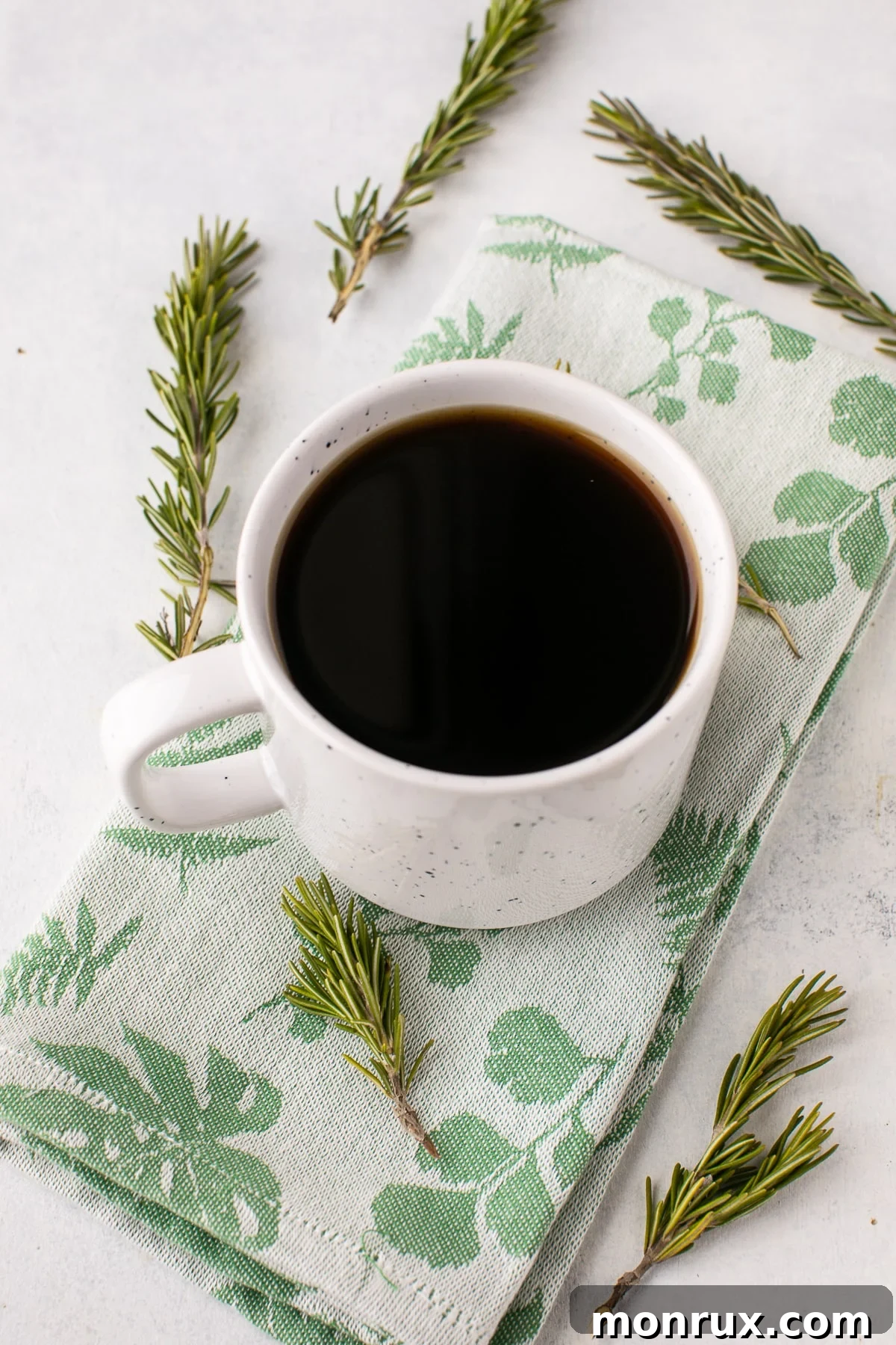 A crisp white mug filled with steaming Rosemary Coffee, adorned with fresh rosemary sprigs on a rustic wooden table, suggesting a healthful morning ritual.