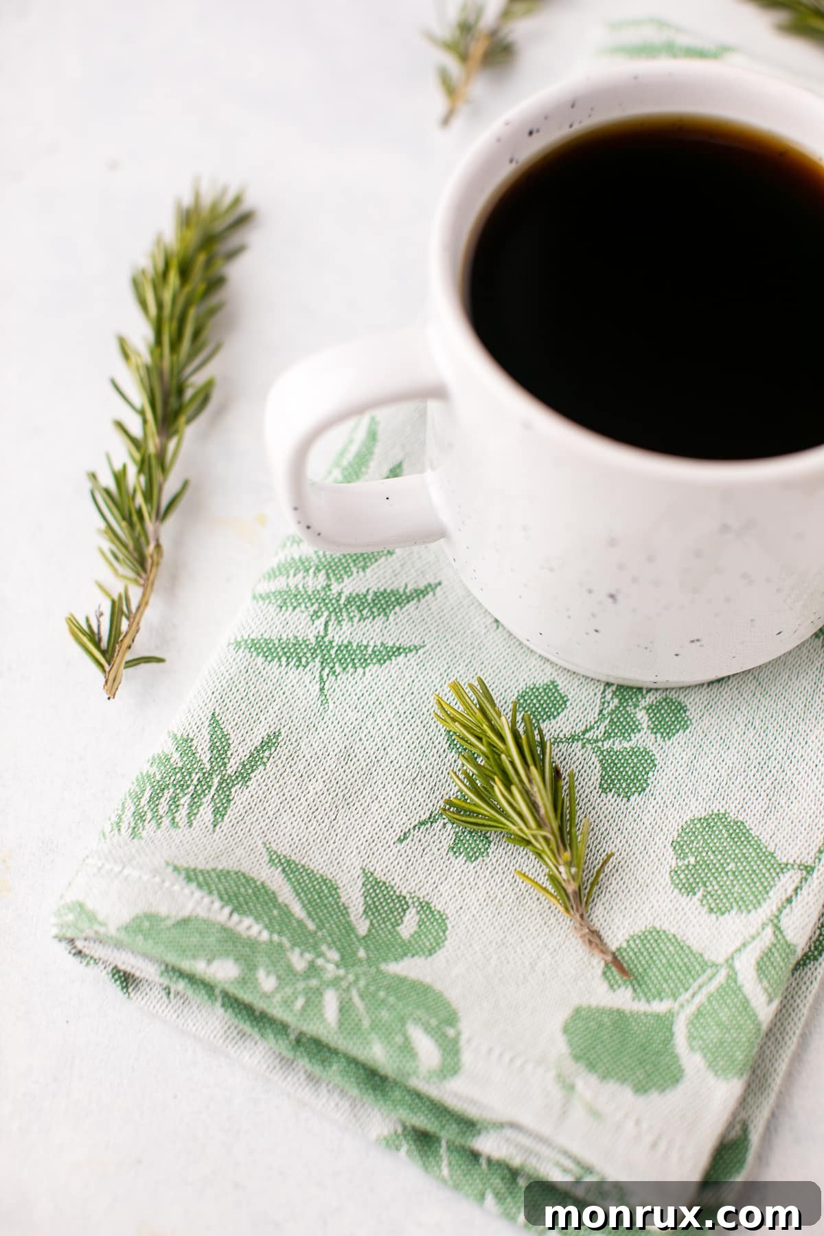 A stylish cup of freshly brewed Rosemary Coffee, garnished with several vibrant sprigs of fresh rosemary, positioned on a sleek surface.