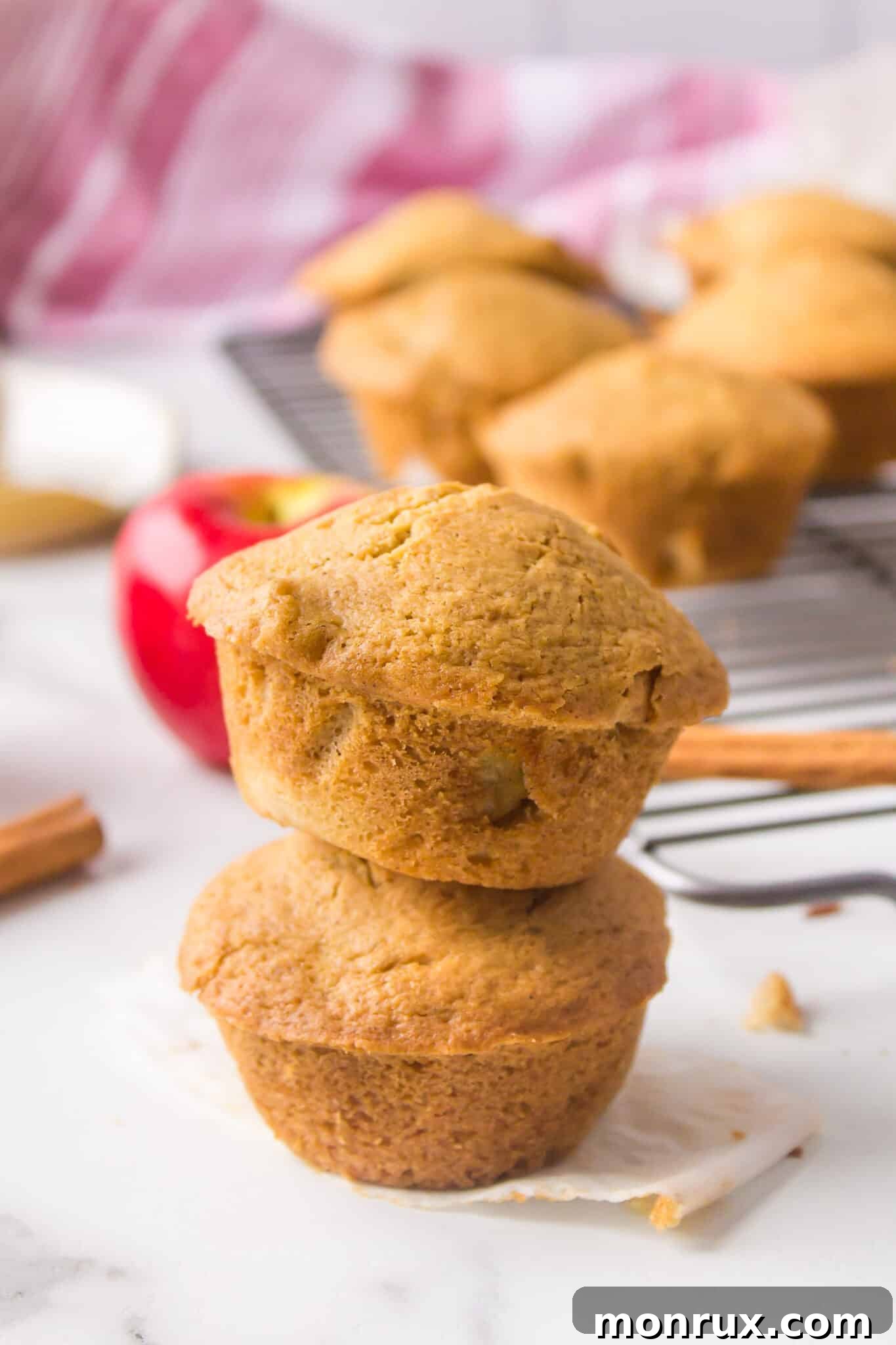 Two golden-brown gluten-free apple muffins, stacked on a rustic kitchen counter with sliced apples in the background.