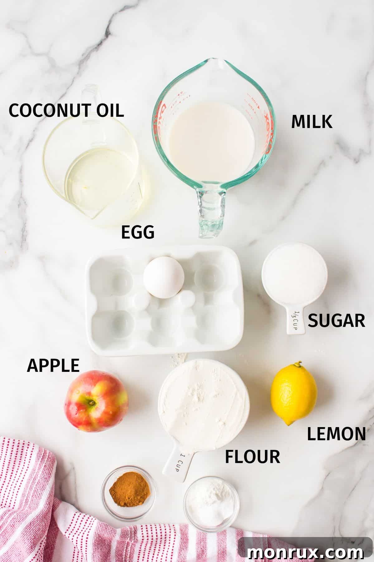 A collection of ingredients laid out on a white surface, including diced apples, gluten-free flour, milk, sugar, oil, and spices, ready to make gluten-free apple muffins.