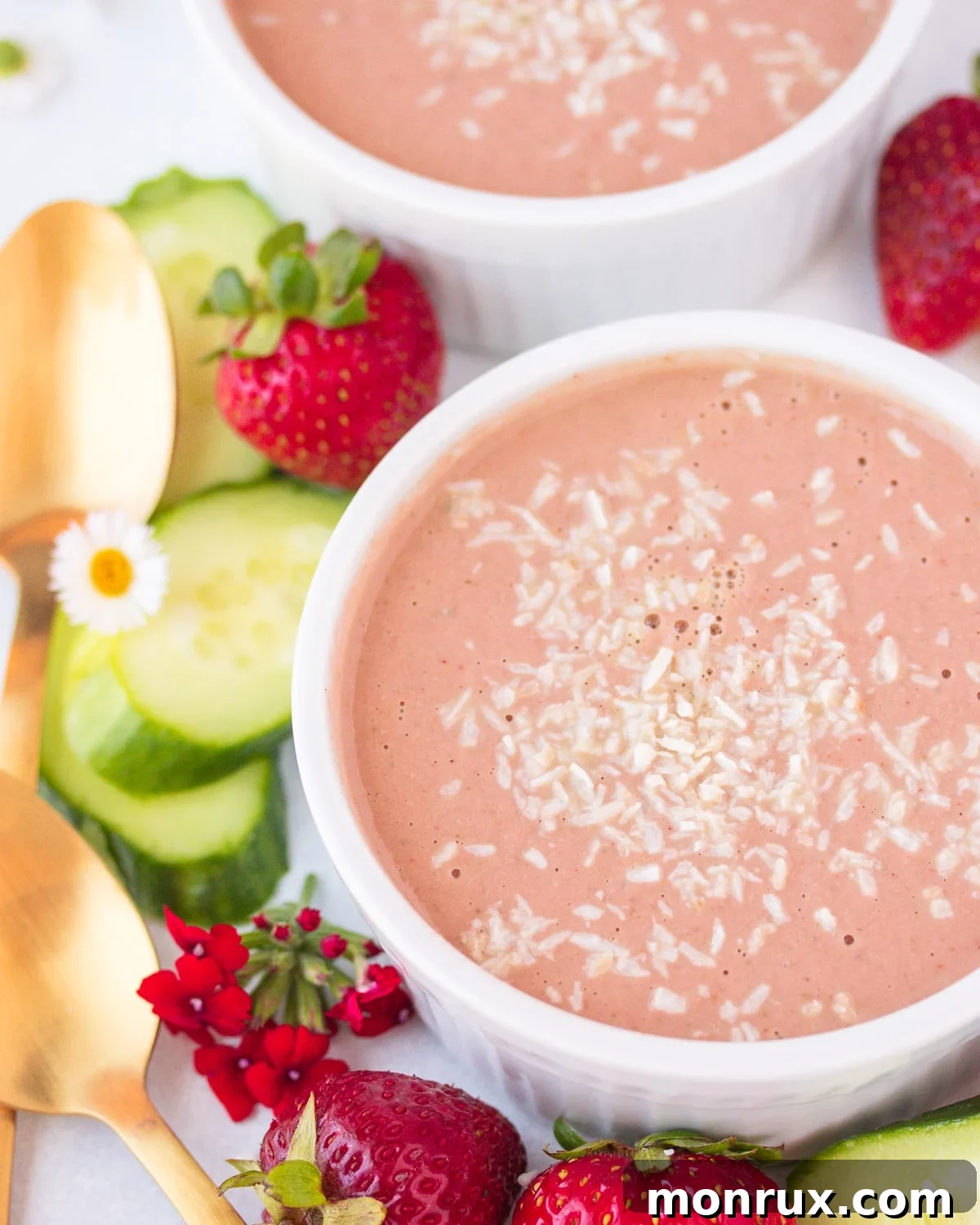 Two bowls of cold strawberry bisque soup, garnished with shredded coconut, set on a rustic wooden surface. The vibrant pink hue of the soup contrasts beautifully with the white coconut.