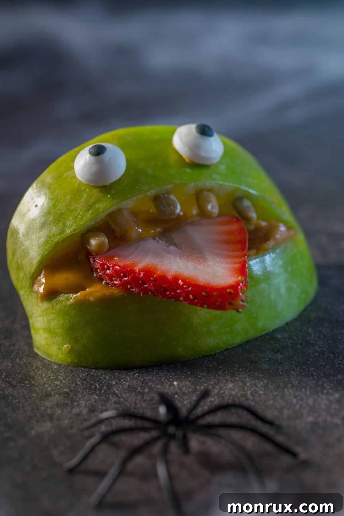 A humorous face crafted from an apple wedge and strawberry slice, with capers for teeth and candy eyes.