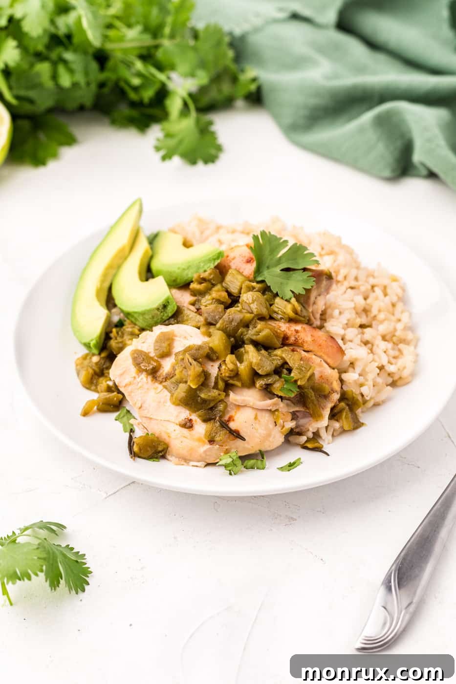 A vibrant bowl of Slow Cooker Green Chile Chicken, served with a side of rice, sliced avocado, and fresh cilantro, on a rustic wooden table.