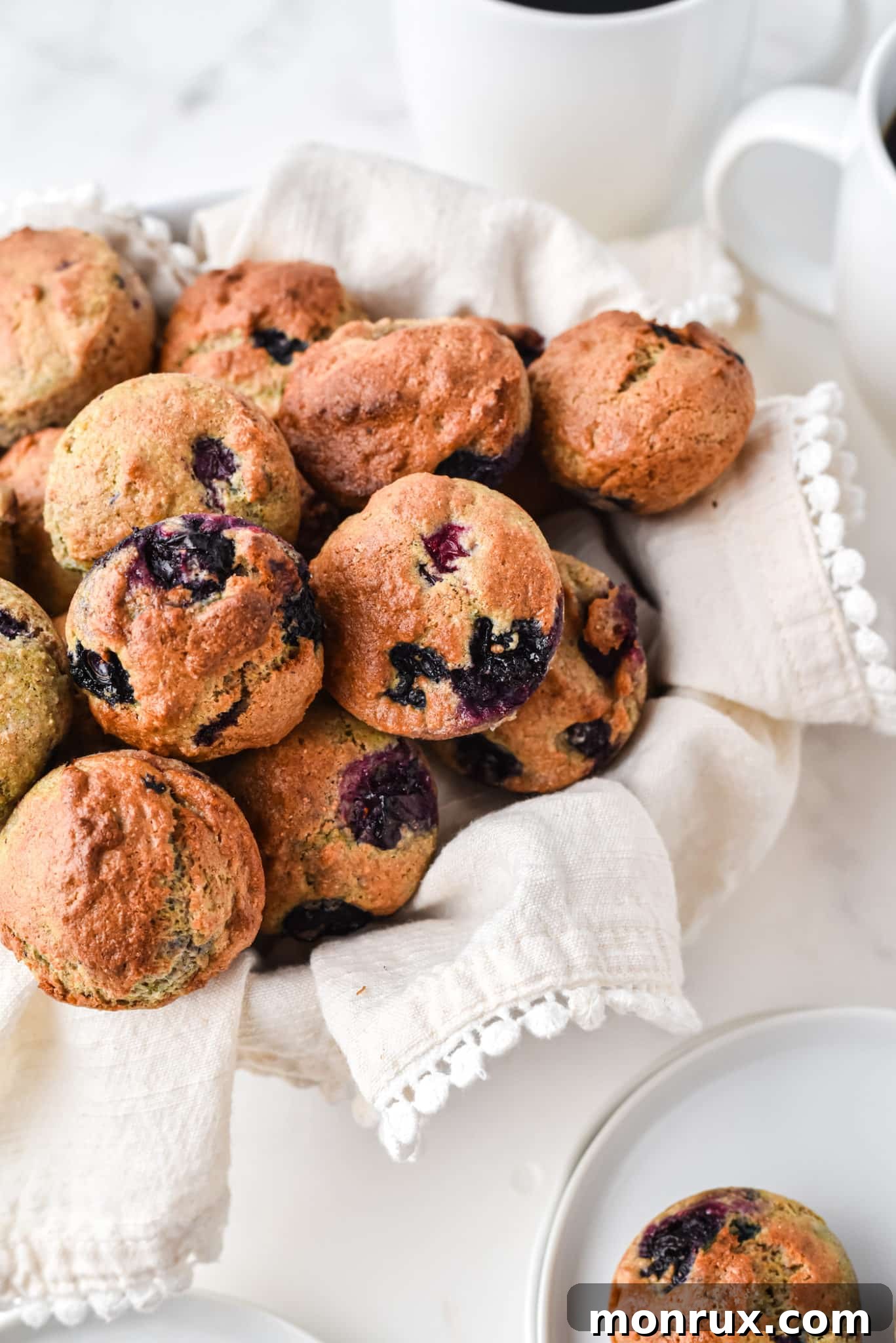 A basket filled with freshly baked mini blueberry muffins, lined with a striped towel, next to a steaming mug of coffee, evoking a cozy breakfast scene.