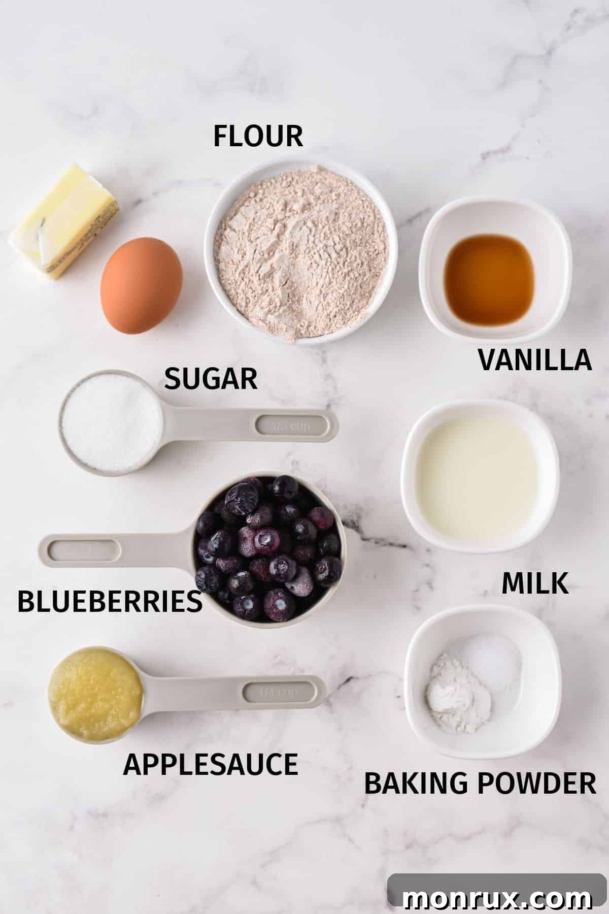 Bowls of measured ingredients for blueberry muffins laid out on a clean white surface, showcasing flour, sugar, blueberries, and other components ready for mixing.