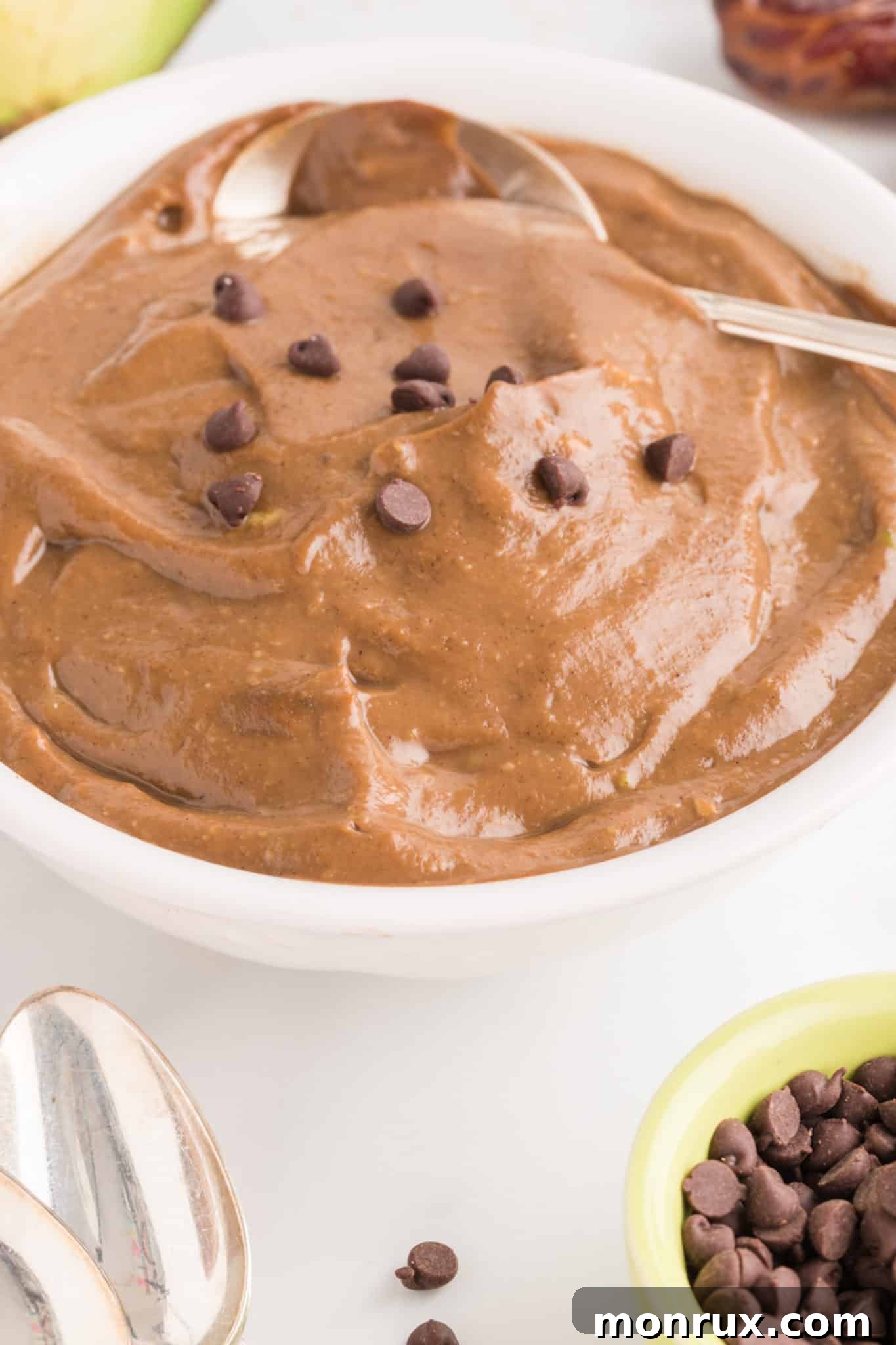 A white bowl with a spoon is filled with creamy, dark avocado chocolate pudding topped generously with rich chocolate chips. The image evokes a sense of indulgence and healthy dessert pleasure.