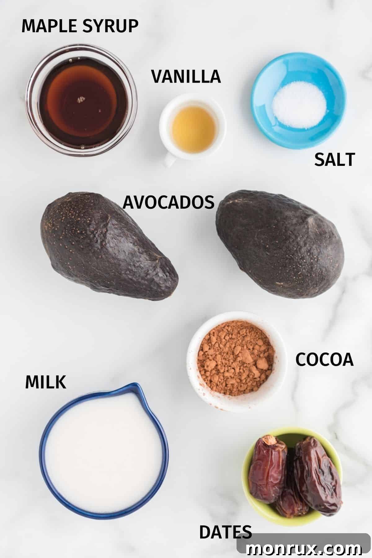 Various ingredients for avocado chocolate pudding are neatly arranged on a pristine white surface, including ripe avocados, cocoa powder, dates, and maple syrup, ready for preparation.