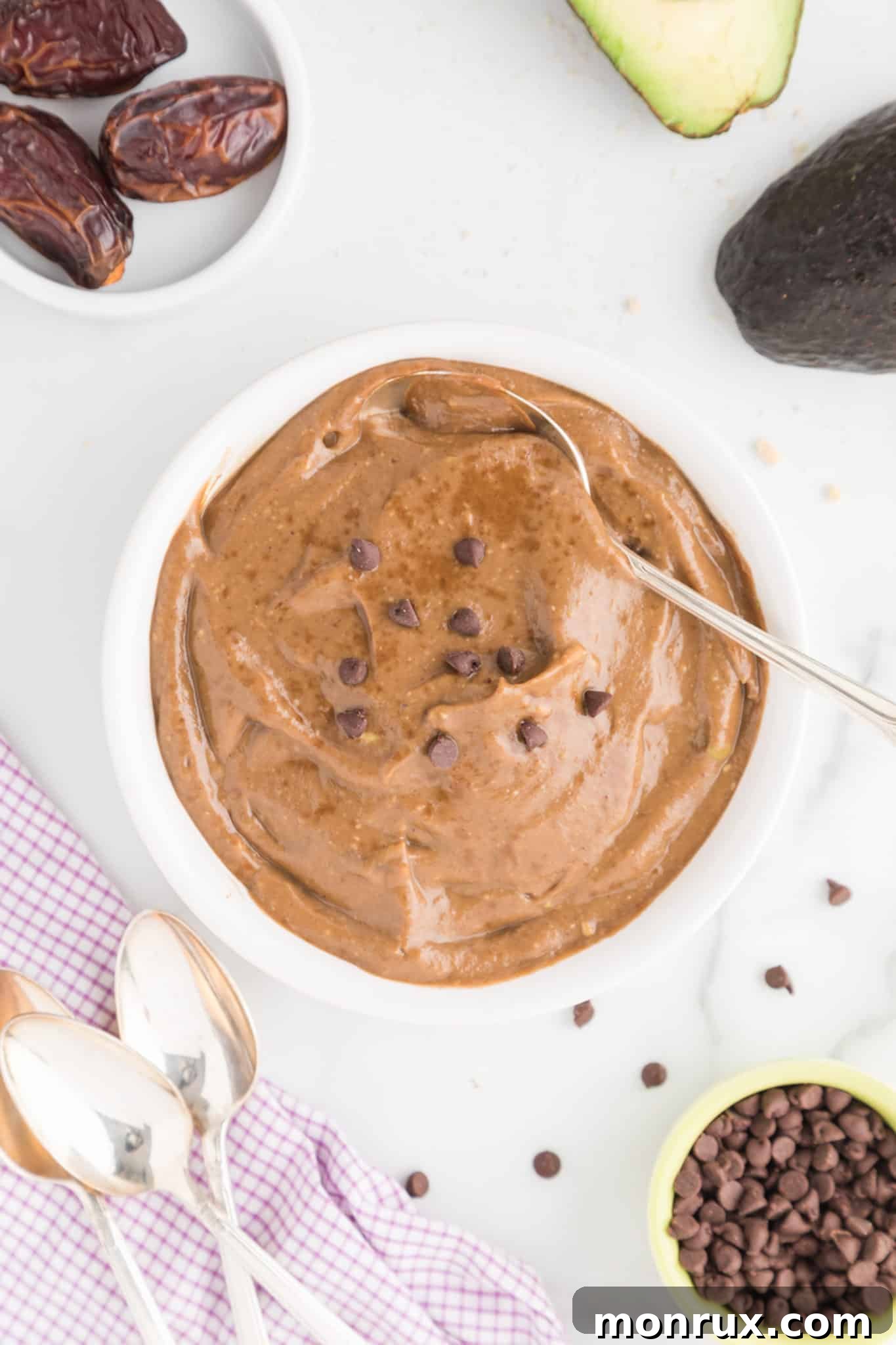 A top-down view of a white bowl filled with dark, creamy avocado chocolate pudding, adorned with chocolate chips and a spoon resting beside it, highlighting its inviting texture.