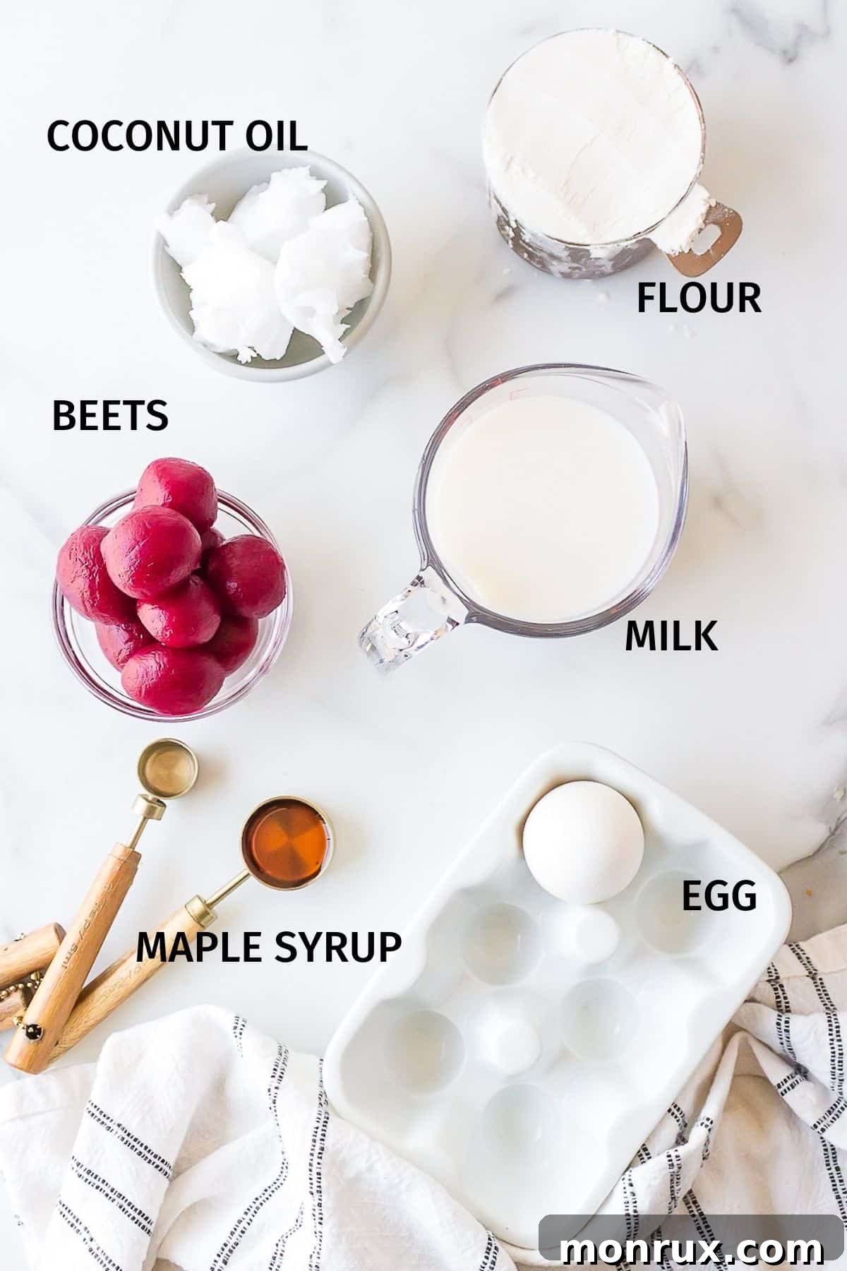 Individual bowls containing pink waffle ingredients: almond milk, an egg, melted coconut oil, maple syrup, all-purpose flour, baking powder, and roasted beets, arranged neatly on a white surface.