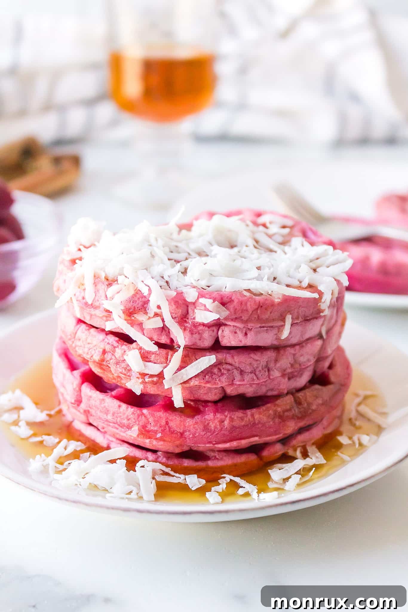 A stunning stack of pink waffles, generously topped with shredded coconut and a cascade of golden maple syrup, beautifully presented on a pristine white plate.