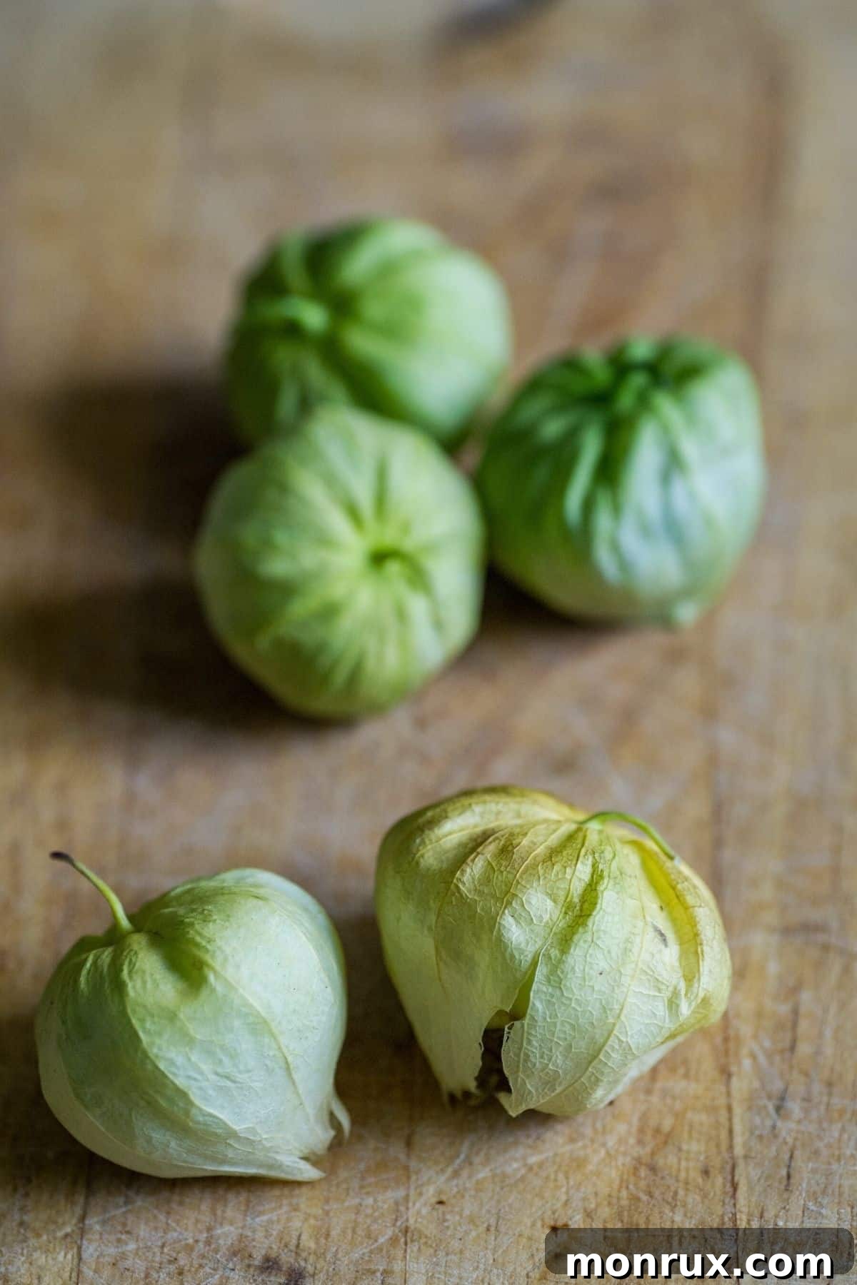 Several fresh tomatillos with their papery husks partially peeled back, revealing the green fruit on a wooden countertop.
