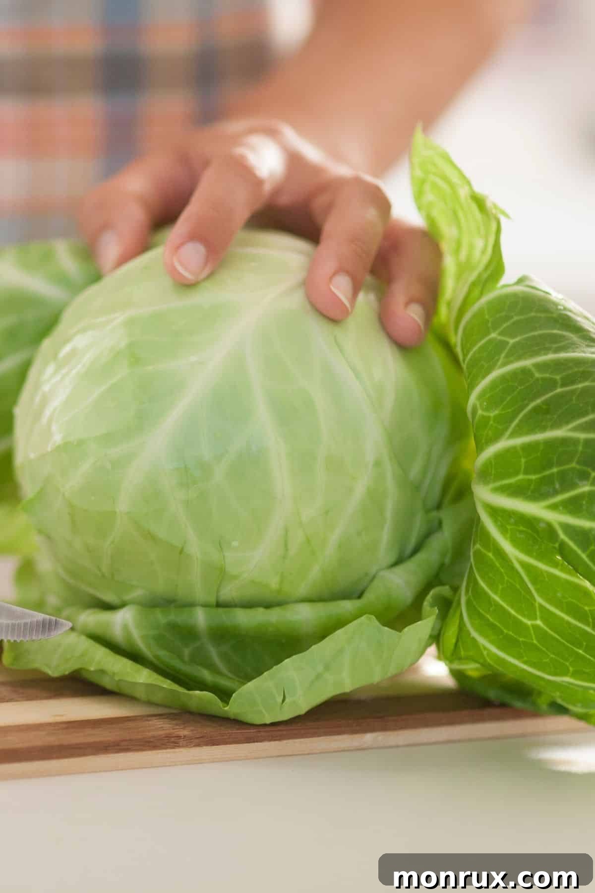 A person's hands cutting a large head of green cabbage on a wooden board, showing its dense layers.
