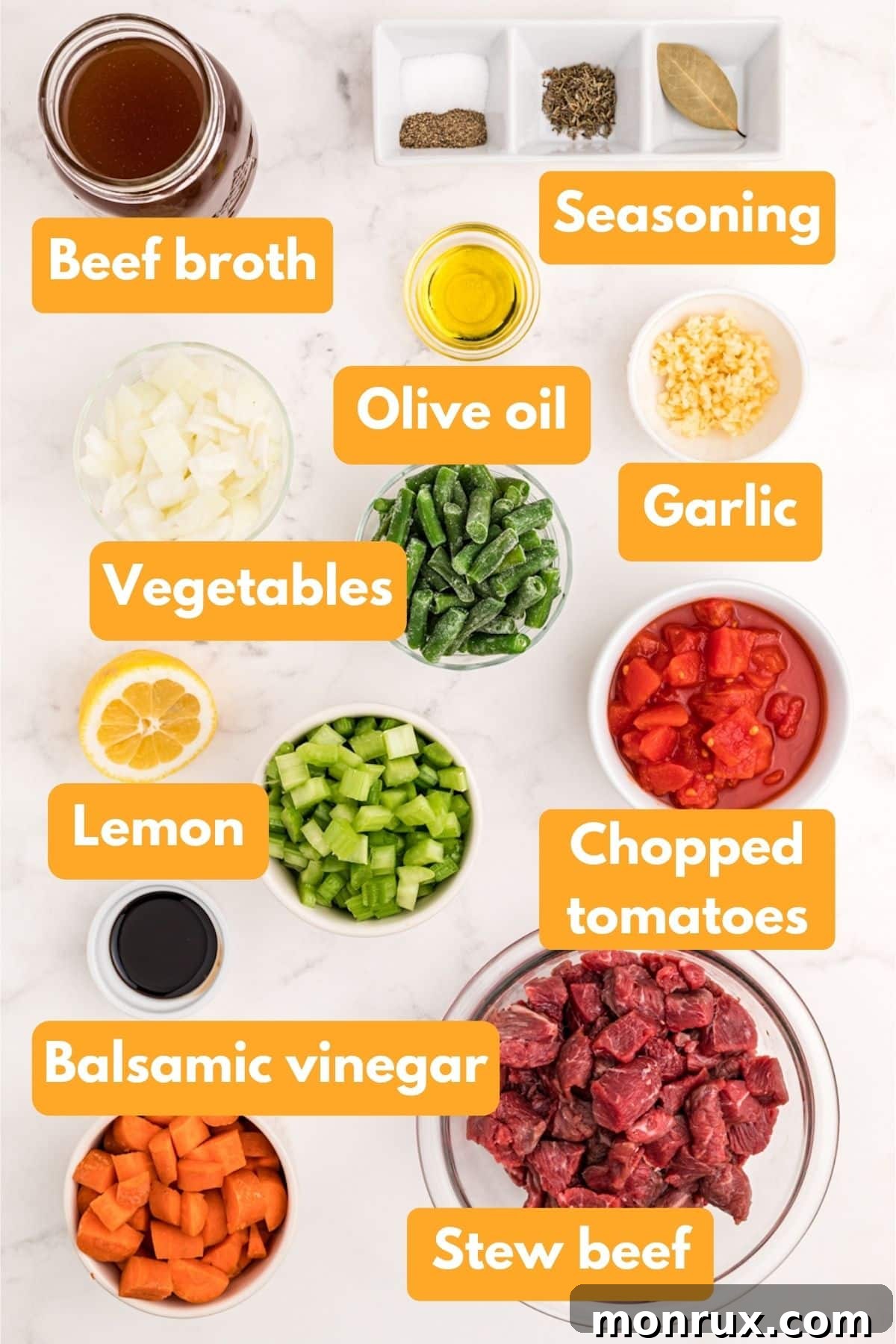 A vibrant selection of fresh ingredients including chunks of stew beef, chopped onions, carrots, celery, garlic, beef broth, balsamic vinegar, and a lemon, laid out on a cutting board.