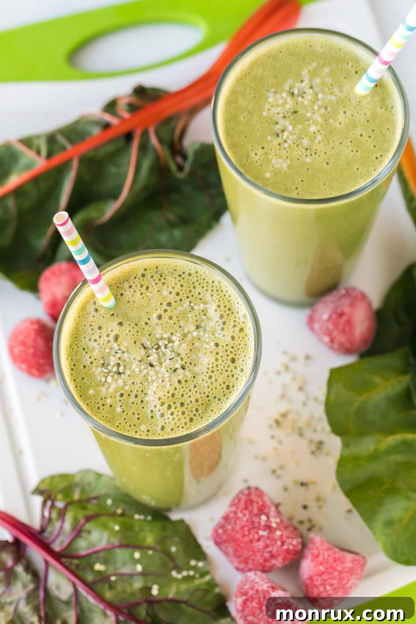 Two glasses of vibrant green Swiss chard smoothie, garnished with fresh berries and a straw, sitting on a rustic wooden table.