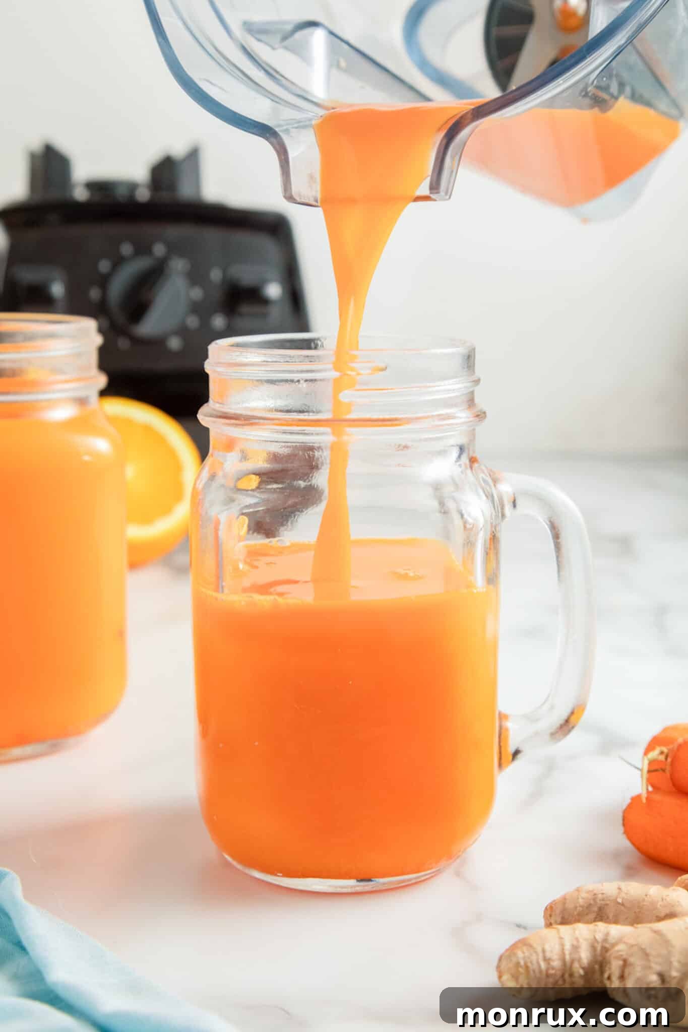 A hand pouring fresh carrot juice into a tall glass, with mason jar mugs in the background.