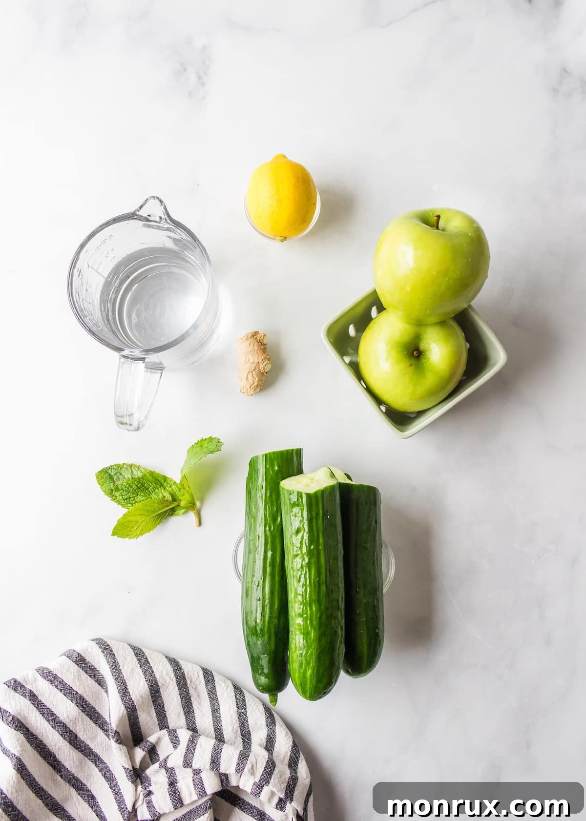 Fresh ingredients for cucumber juice laid out on a table: whole cucumbers, green apples, a lemon, ginger root, and mint leaves.