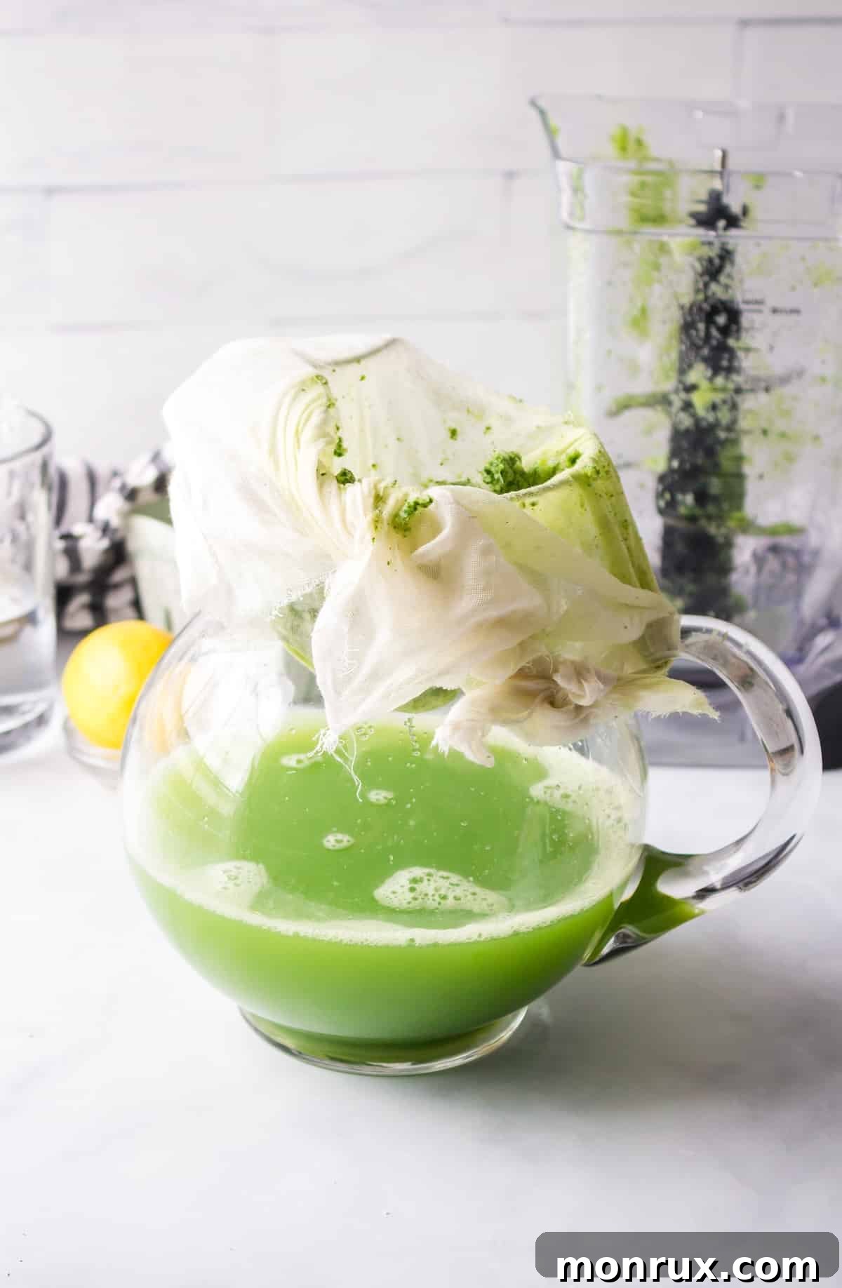 Green juice being strained through a nut milk bag into a glass pitcher, separating the liquid from the pulp.