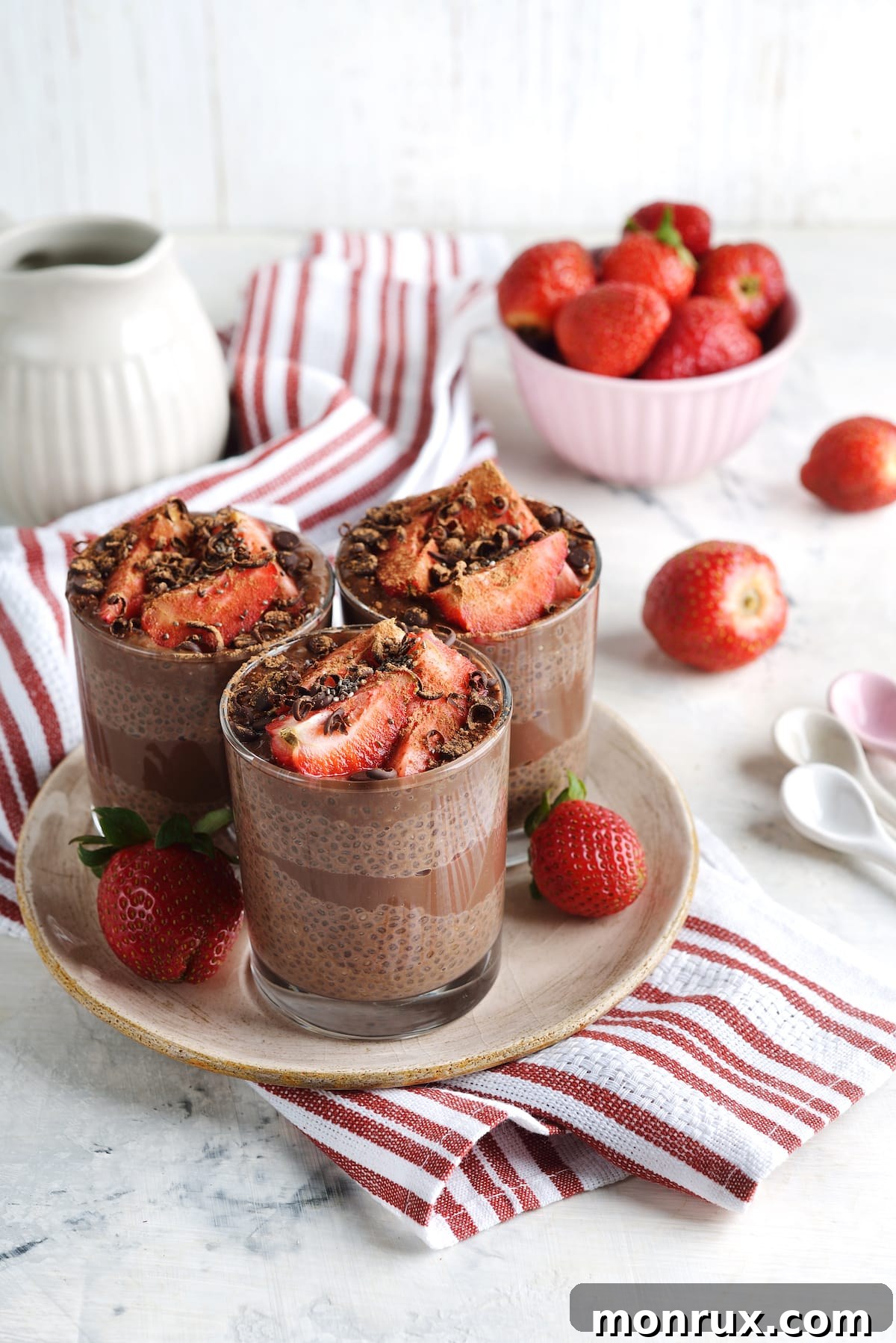 Three glass jars of strawberry chocolate chia pudding, garnished with fresh berries and chocolate chips, arranged on a rustic wooden tabletop.