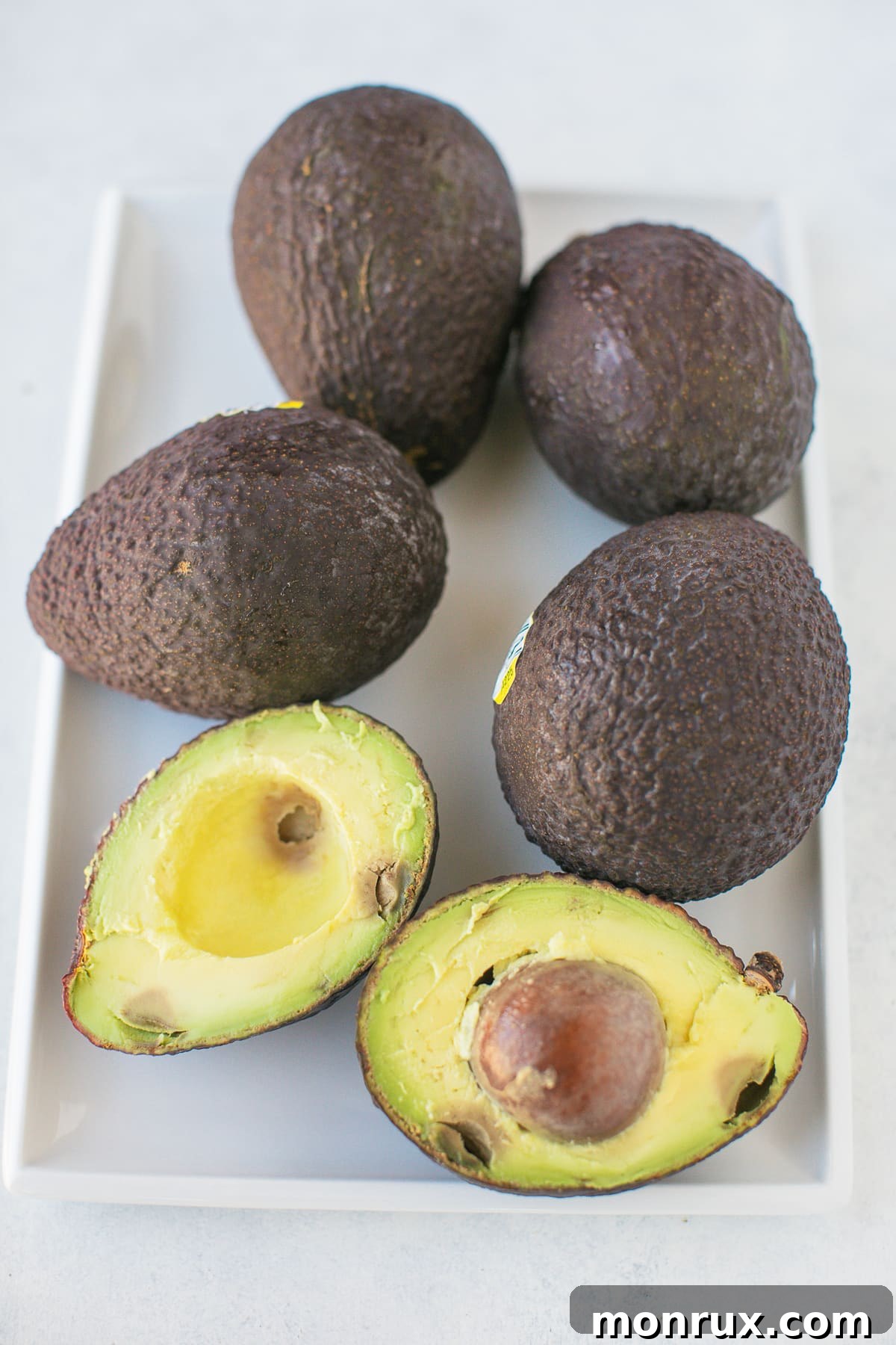 Fresh, ripe avocados on a white platter, with one cut in half, ready for preparation.