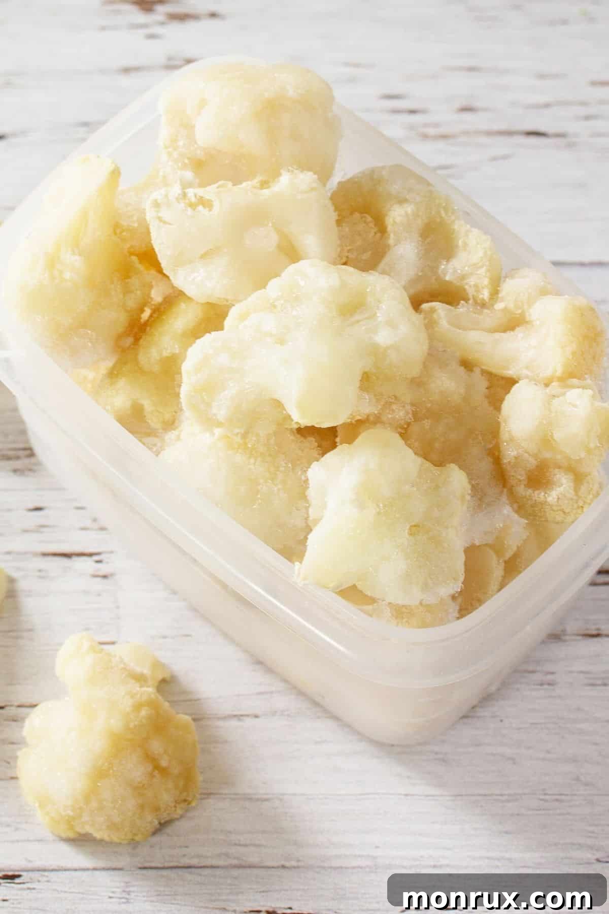 A clear container filled with perfectly frozen cauliflower florets rests on a rustic wooden tabletop, highlighting the freshness and readiness for future use.
