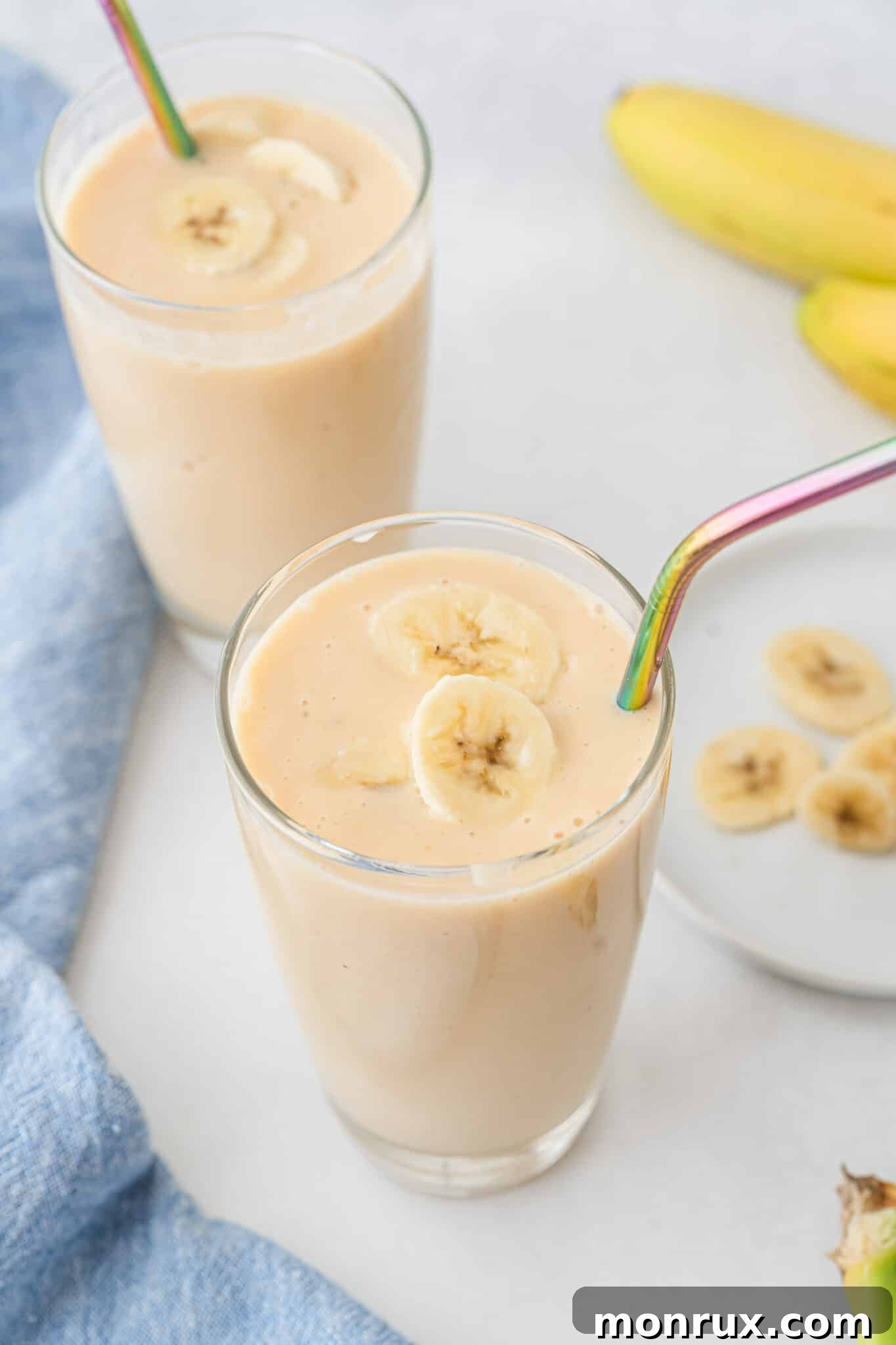 Two glasses of creamy banana peach smoothie with iridescent straws, garnished for a refreshing summer drink.