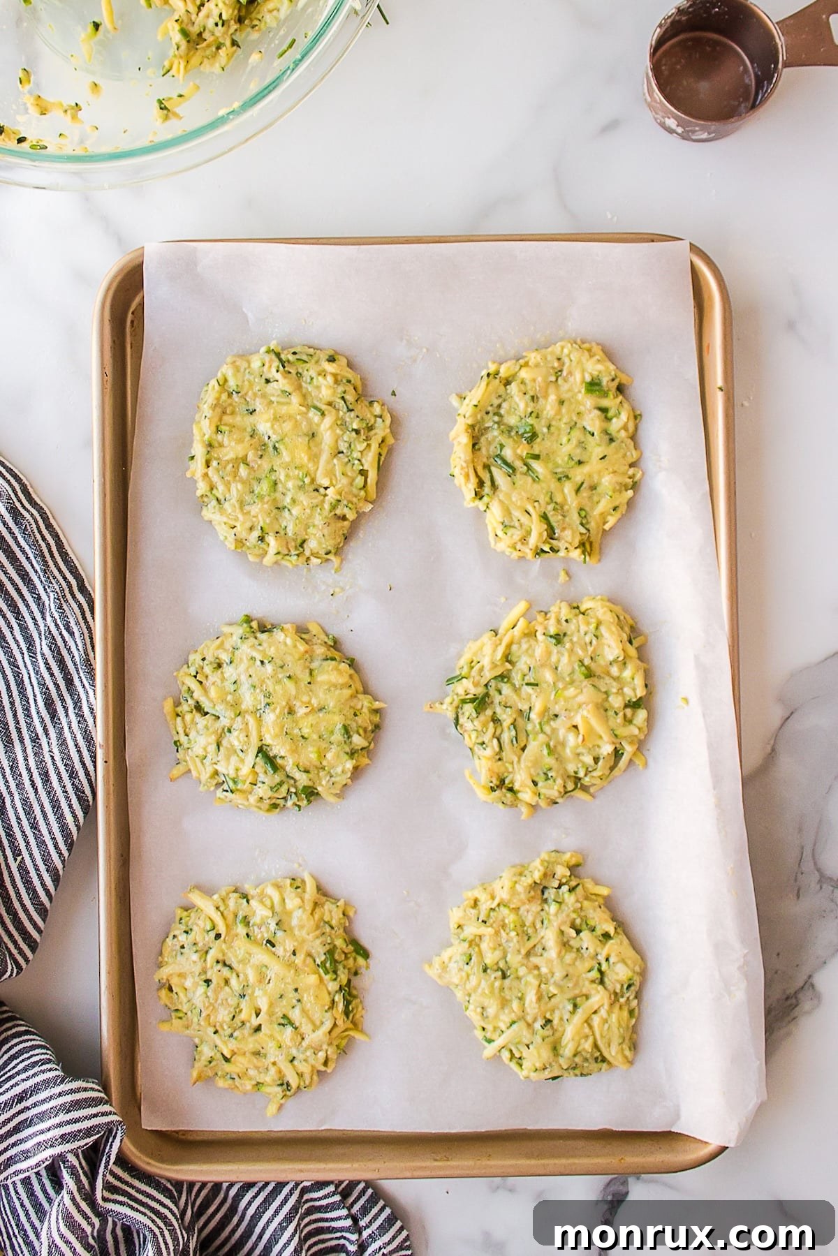 Thin, round zucchini potato fritters neatly arranged on a parchment-lined baking sheet, ready for the oven.