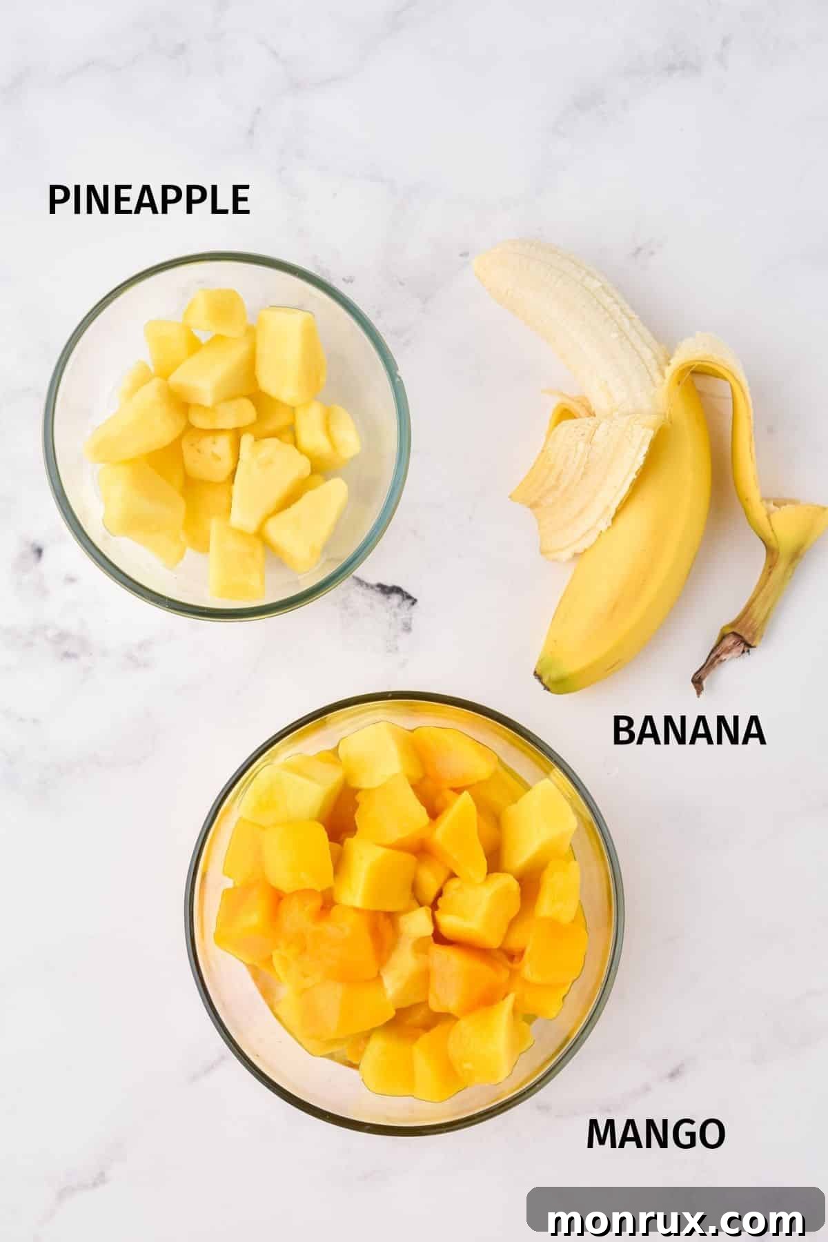 Individual ingredients for mango popsicles, including sliced mango, pineapple chunks, and a banana, displayed on a clean white surface.