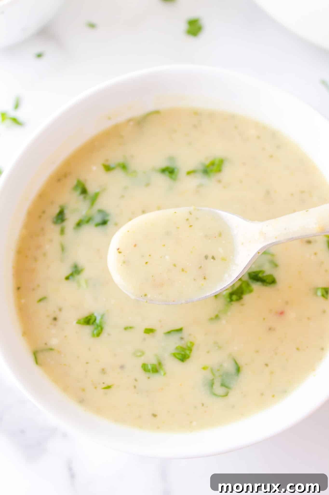 A silver spoon lifting a bite of creamy zucchini potato soup from a pristine white bowl, garnished with fresh herbs.