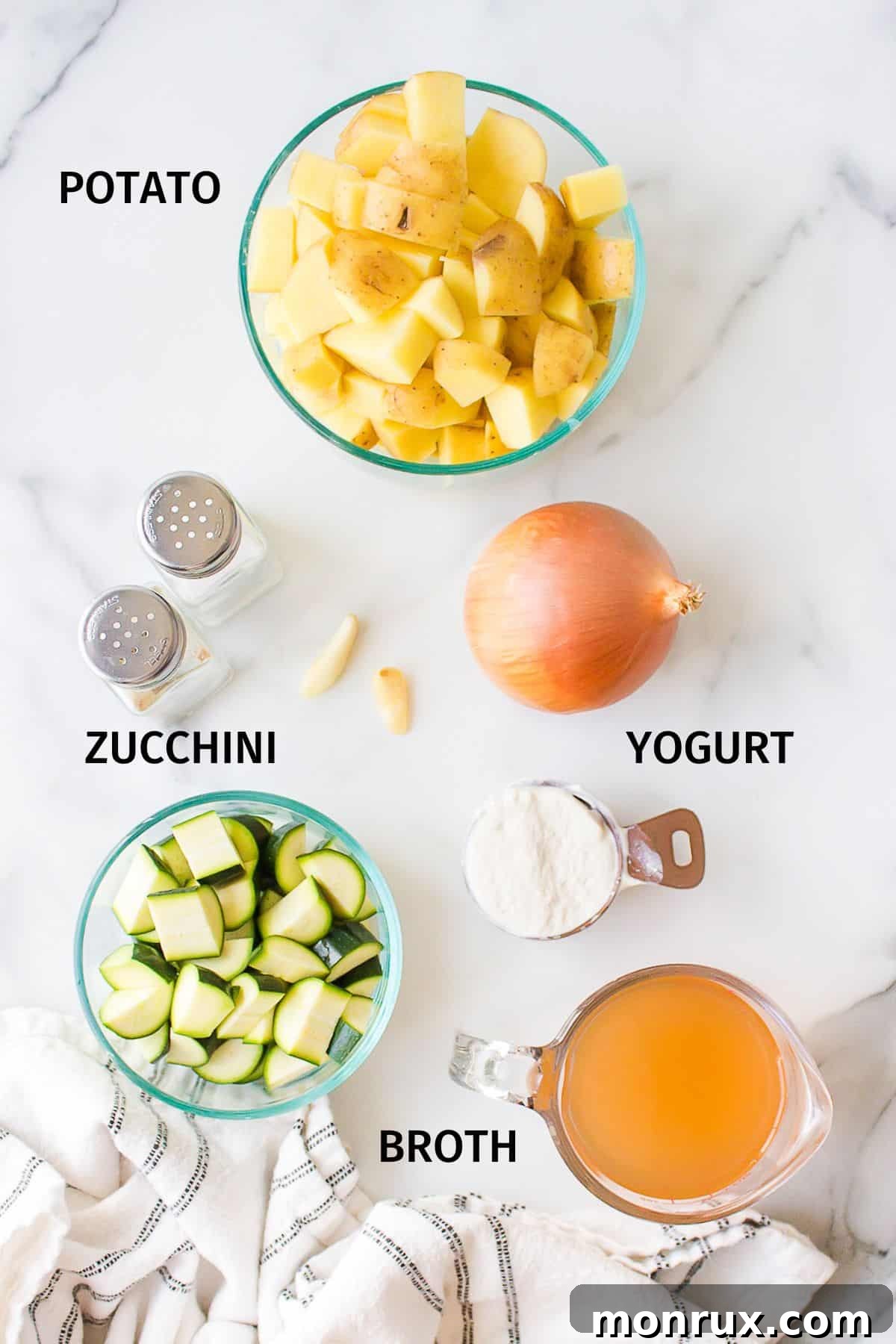 Assorted fresh ingredients for zucchini potato soup, including chopped zucchini, potatoes, onions, and garlic, arranged in small bowls on a white countertop.