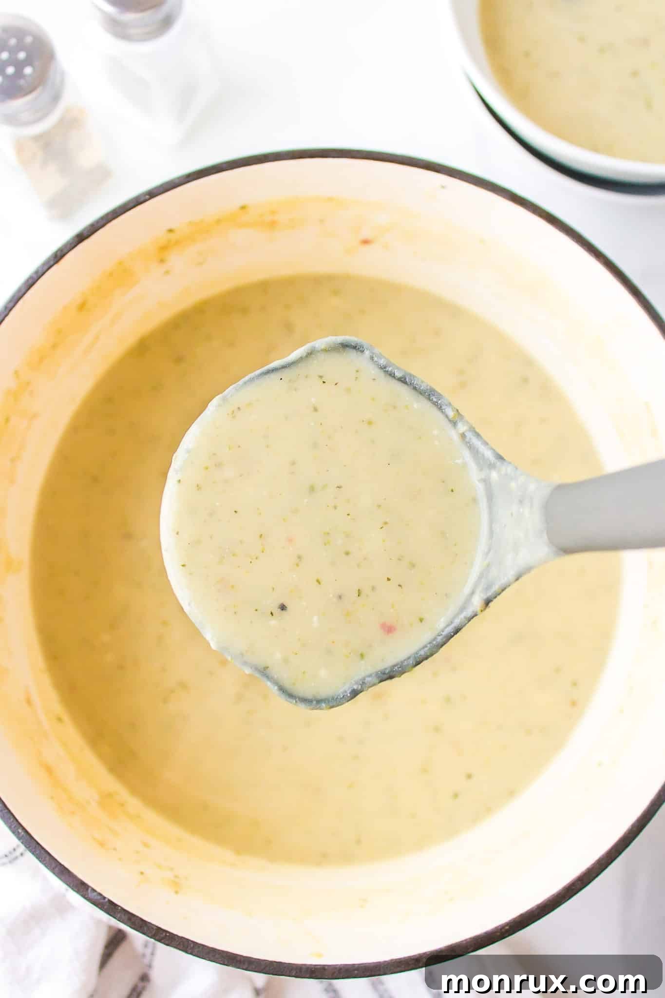 A gray ladle filled with creamy zucchini potato soup is held above a steaming Dutch oven, invitingly ready to serve.