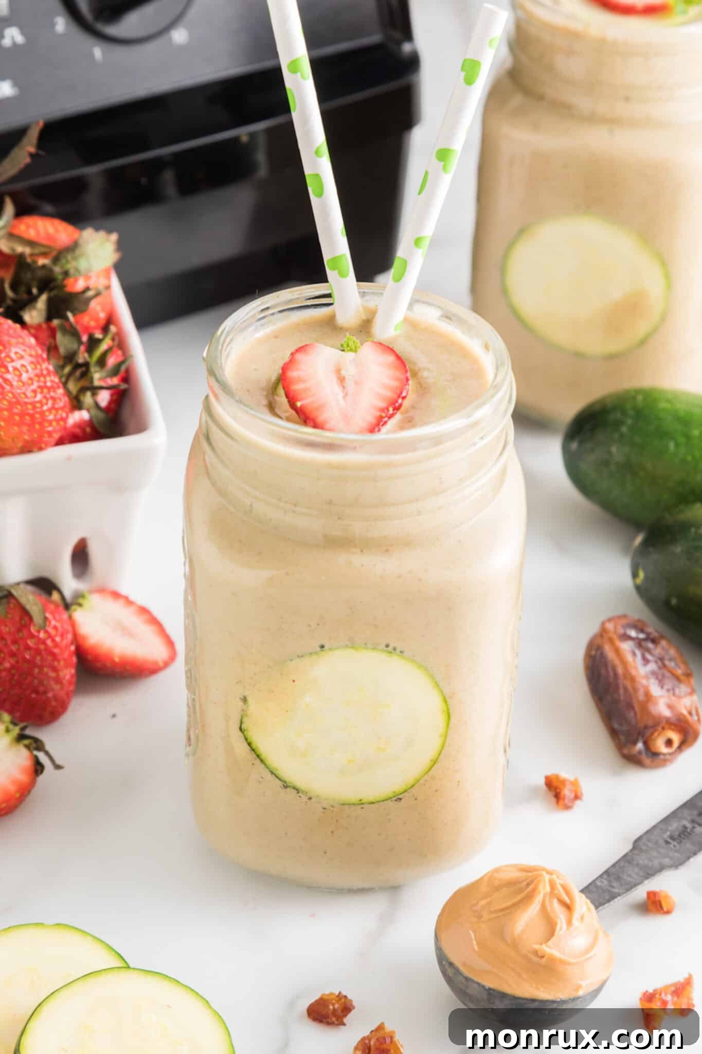 A vibrant strawberry zucchini smoothie in a mason jar, garnished with fresh strawberries and two straws, on a light background.