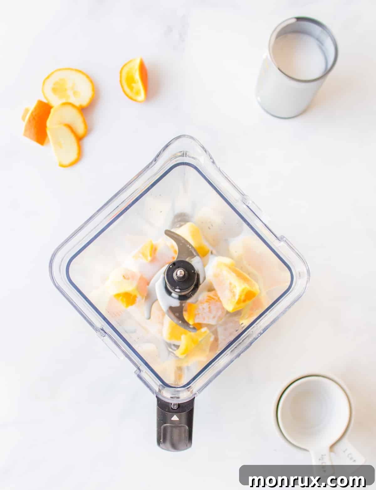 A blender filled with coconut milk, peeled oranges, and frozen pineapple, ready to be blended.