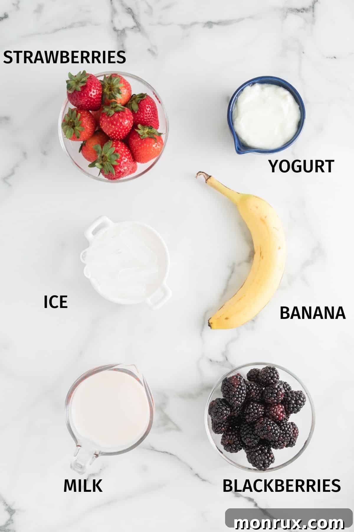 Small bowls containing fresh strawberries, blackberries, a sliced banana, and a glass of milk, laid out on a white surface, ready for smoothie preparation.